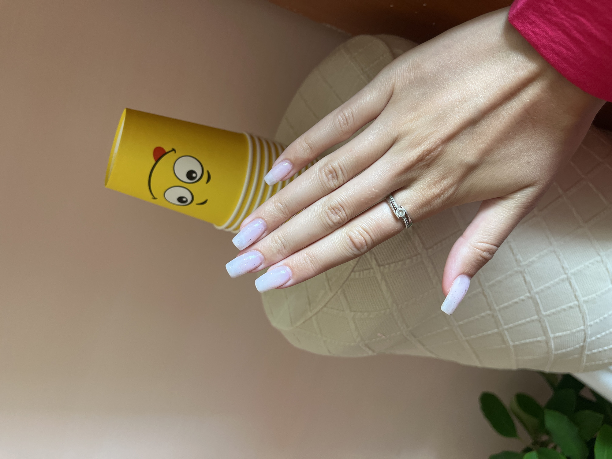 A close-up shot showcases a woman's hand adorned with a ring and meticulously manicured nails. The nails are long and square-shaped, painted in a subtle, iridescent pinkish-white hue, with fine glitter embedded in the polish. On the ring finger, a delicate silver band is visible, featuring a small, clear stone and a row of tiny sparkling diamonds.
The hand rests on a textured, pale cream-colored surface, possibly a pillow or fabric, which provides a soft contrast to the skin tone and nails. To the left of the hand, a bright yellow paper cup is positioned. The cup is decorated with a whimsical cartoon face, complete with large, wide eyes, a cheeky red tongue sticking out, and simple black outlines for the mouth and eyebrows.
The background is a soft, muted pinkish-brown, suggesting an indoor setting with diffused lighting. The overall ambiance of the image is calm and visually appealing, focusing on the details of the hand, the nails, and the playful cup. No specific time of day or weather conditions can be inferred. The location context of Novoaltaysk, Russia, does not provide any visible cues within the image itself. There is no visible text in the image.