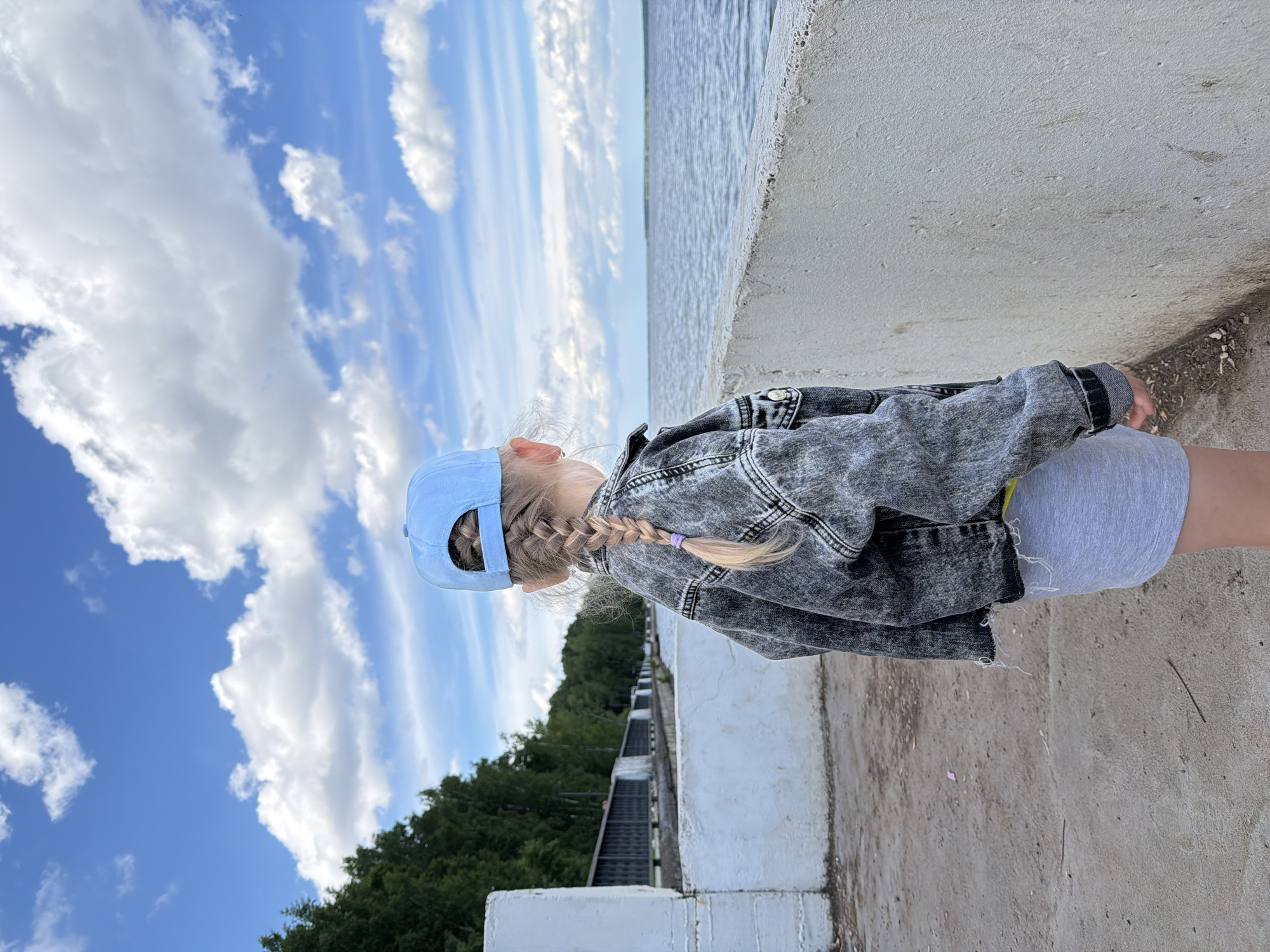 A young girl, viewed from behind, stands on a concrete embankment overlooking a body of water. She is wearing a light blue baseball cap, a grey denim jacket, and a grey skirt. Her blonde hair is braided in a single plait. The background shows a line of trees and a cloudy sky. The scene appears to be along the Kama River embankment in the city of Chaikovsky, Russia.