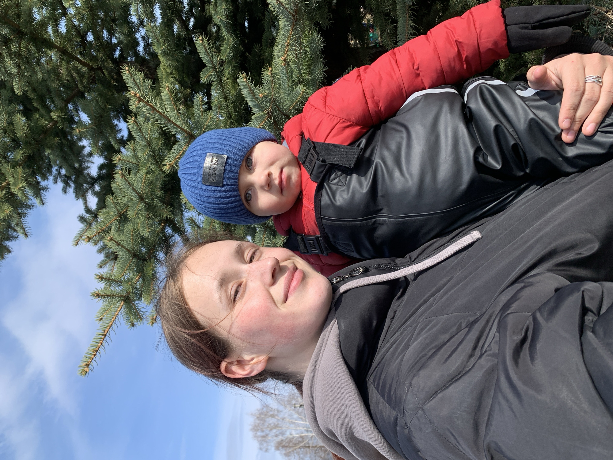 A young woman and a baby are captured in a close-up, rotated selfie taken outdoors in Фоки, Russia. The woman, smiling gently with light skin and dark hair, wears a dark quilted jacket over a grey hooded sweatshirt. A silver-colored ring is visible on her left ring finger. She holds the baby, who wears a blue knit beanie featuring a black patch with the text "ENJOY THE NOW EVERY DAY", a red puffy jacket, and a black snowsuit. The baby looks towards the camera with wide blue eyes.
The background primarily consists of dense evergreen trees under a bright, clear blue sky with some wispy clouds. Bare deciduous trees are visible further in the distance, indicating a cold season such as winter or early spring. The bright sunlight suggests a sunny daytime setting, contrasting with the warm winter clothing worn by both individuals. The image conveys a warm and affectionate moment between the woman and child, enjoying the outdoors in a natural, forested environment.