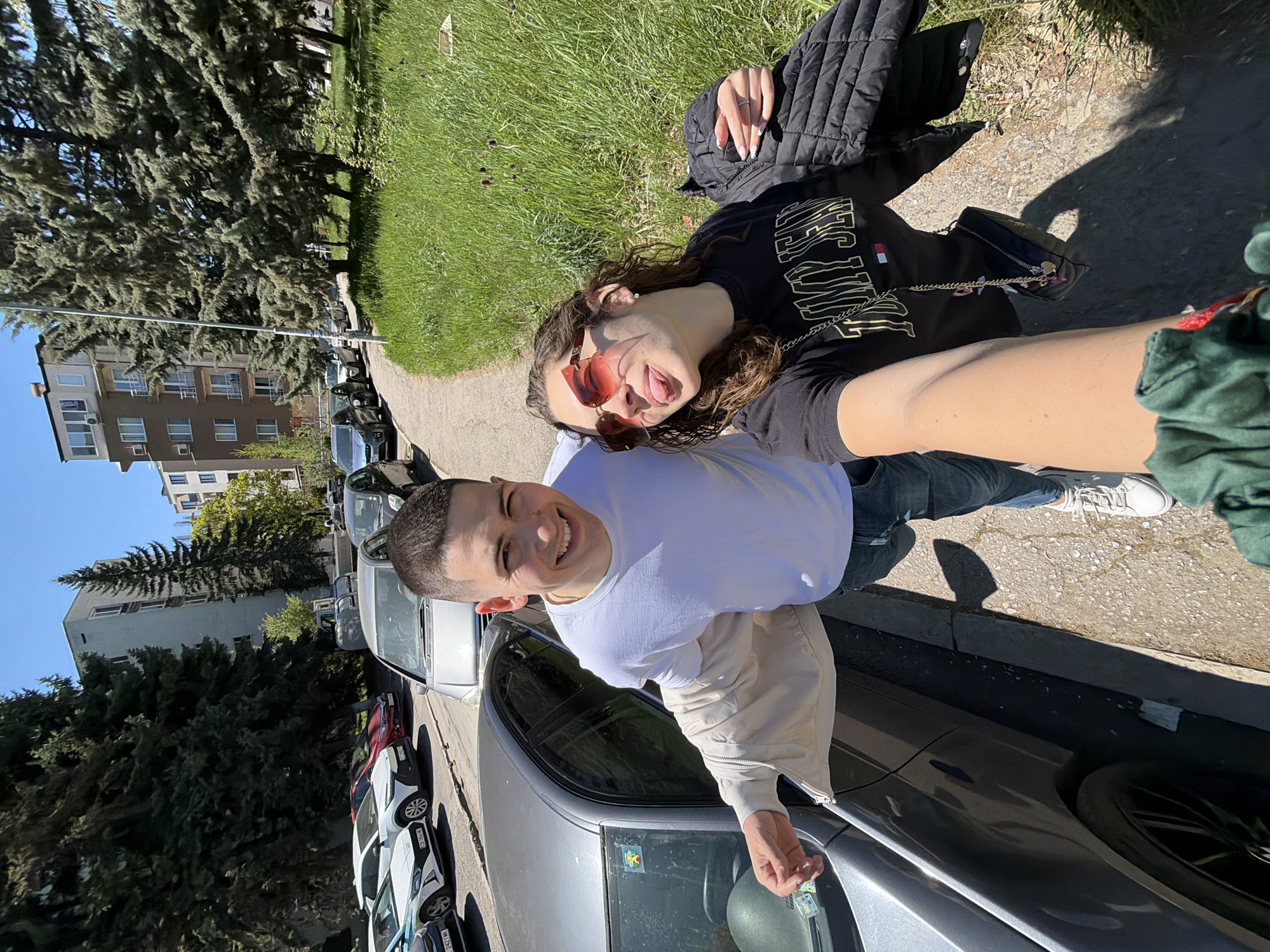 A young man and woman are taking a selfie on a street in Sofia, Bulgaria. The man is standing beside a parked silver car, holding the camera. He is wearing a white t-shirt and a light beige jacket. The woman is lying on the grass beside the sidewalk, wearing a black shirt with gold lettering, red sunglasses, and a black quilted jacket. She is sticking her tongue out. The background shows a residential street with parked cars, trees, a sidewalk, and apartment buildings.