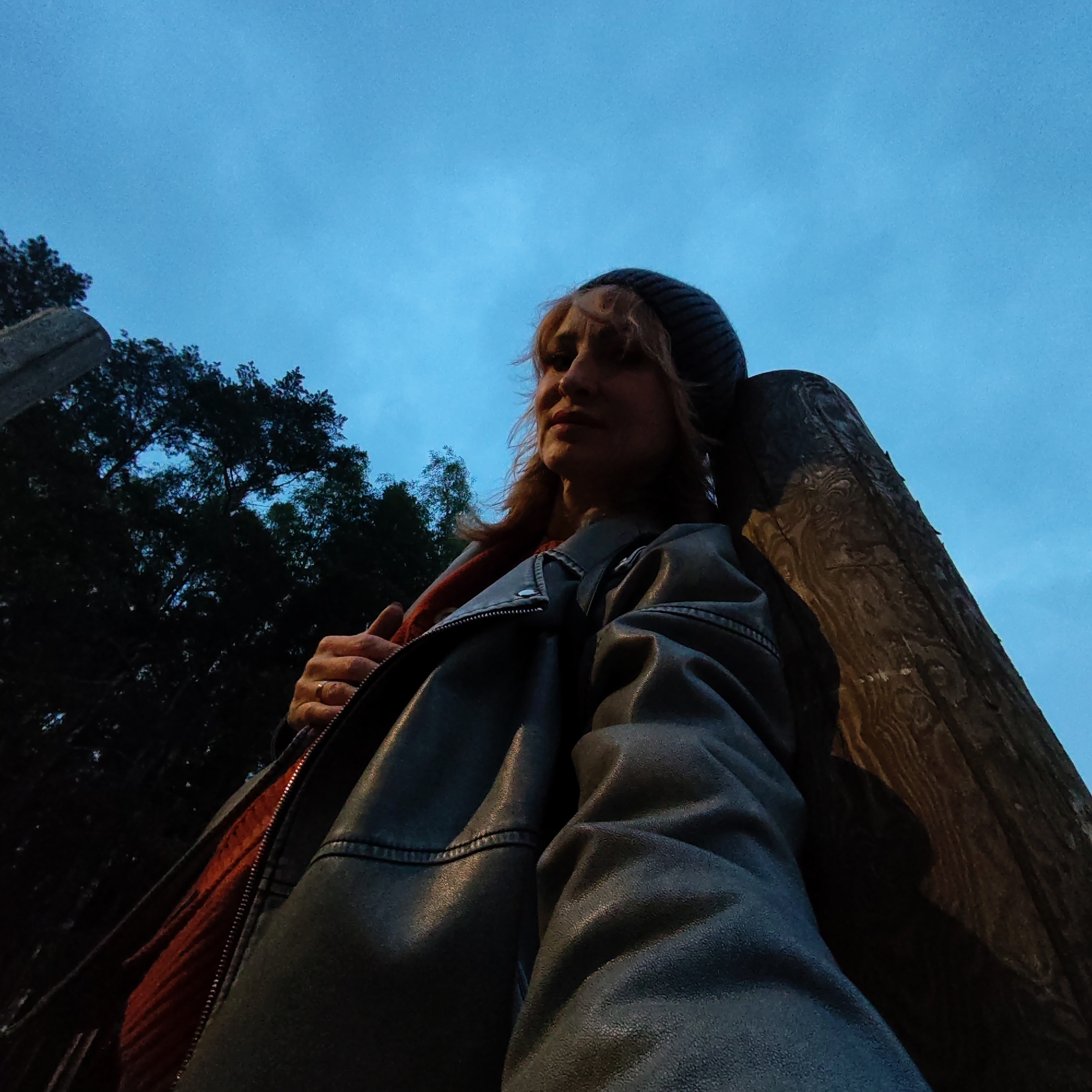 A low-angle shot shows a woman leaning against a wooden post in Chaikovsky, Russia. She is wearing a dark beanie, a gray leather jacket with a partially visible red lining, and a wedding ring. In the background are trees and a blue, overcast sky. The wooden post has a dark, textured surface.