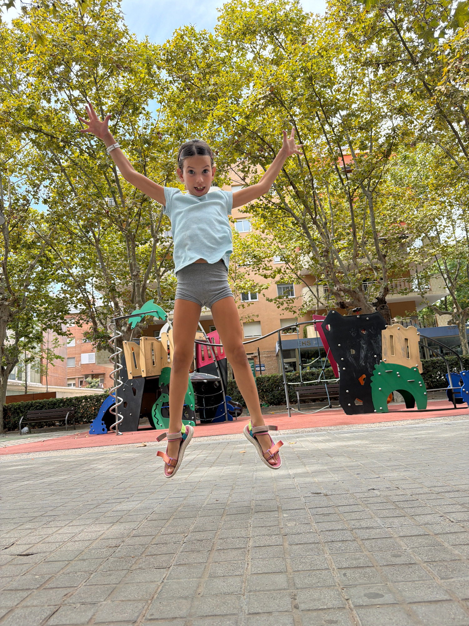 A young girl with dark hair pulled back in a braid is captured mid-air, jumping with her arms outstretched in a playground. She wears a light blue t-shirt and grey shorts, with pink and white sandals. Her face shows an excited expression, with pink blush applied to her cheeks. The background reveals a colorful playground structure with red, blue, black, and green elements, including slides and climbing frames. Trees with green and yellow leaves provide shade, and buildings with orange facades are visible behind the playground. The ground is paved with grey interlocking bricks. The scene is set in the city of Salou, Spain.