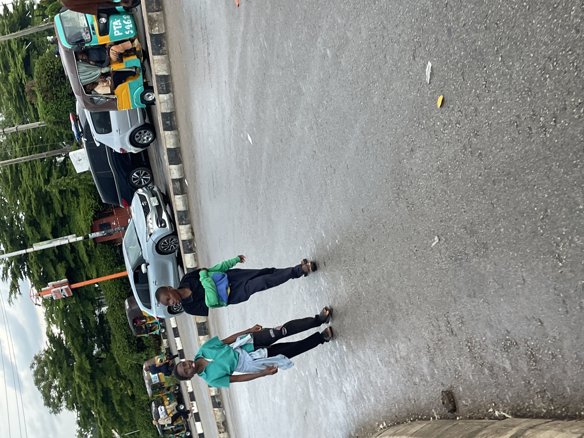 In Jos, Nigeria, two young boys walk on a wet asphalt road. The boy on the left wears a teal polo shirt, black pants, and dark sandals, with a light gray jacket tied around his waist. The boy on the right wears a black t-shirt and dark pants, with a green and blue hooded sweatshirt draped over his shoulders. Behind them, several vehicles are visible: a silver SUV, a black SUV, and two motorized rickshaws, one yellow and turquoise, the other yellow and green. The street is lined with a concrete barrier and a black and white striped curb. Trees and an orange traffic signal with a white sign are visible in the background. The sky is overcast.