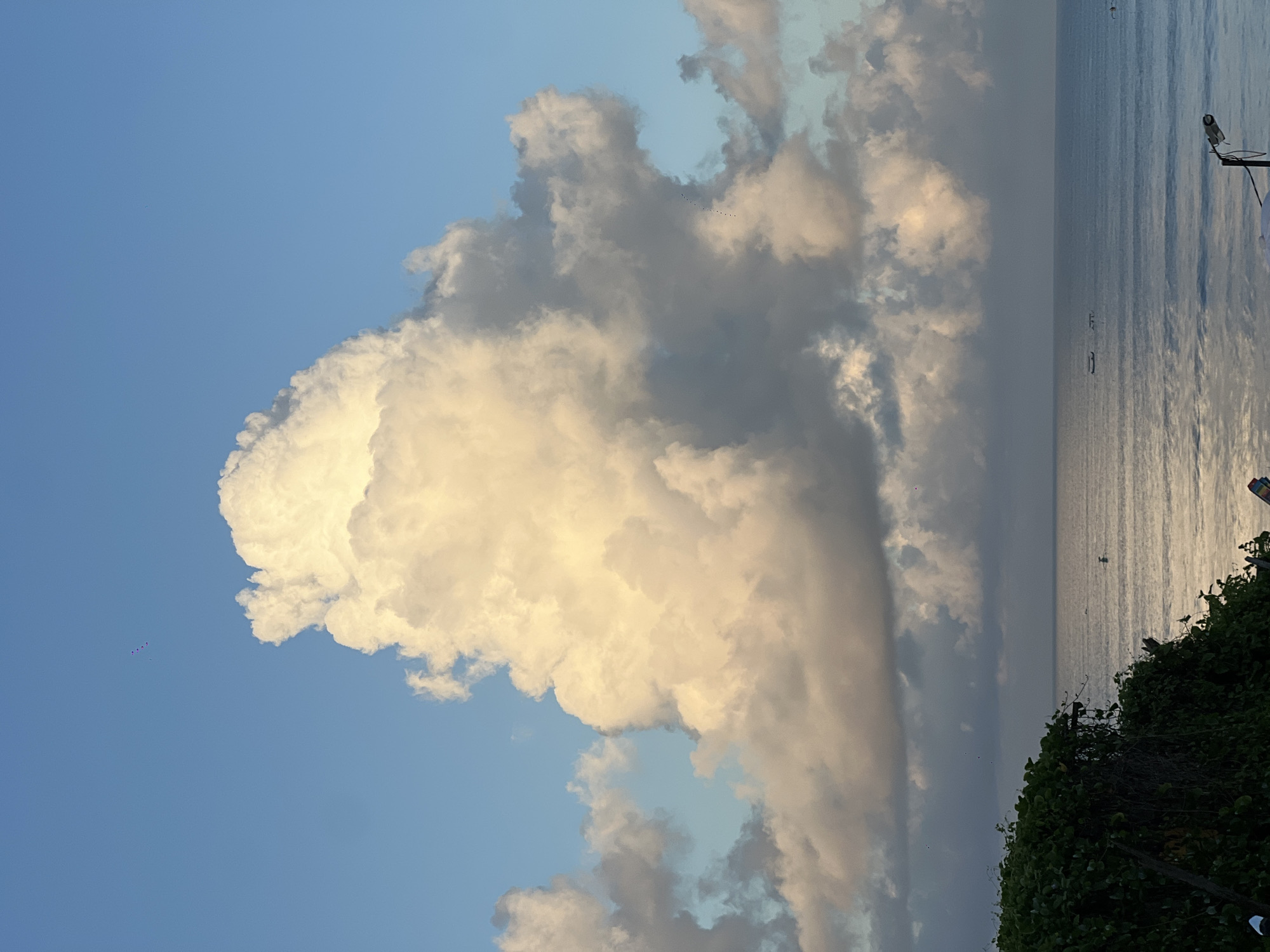 The image shows a large cumulus cloud in the sky. The cloud is white and gray, and it appears to be towering. The sky is blue. To the right, there is an ocean or large body of water with a shimmering surface. The shoreline includes trees or bushes along the bottom right corner of the image. A security camera is affixed on the upper right. There are some birds in the sky. Location: Unknown, Indonesia.