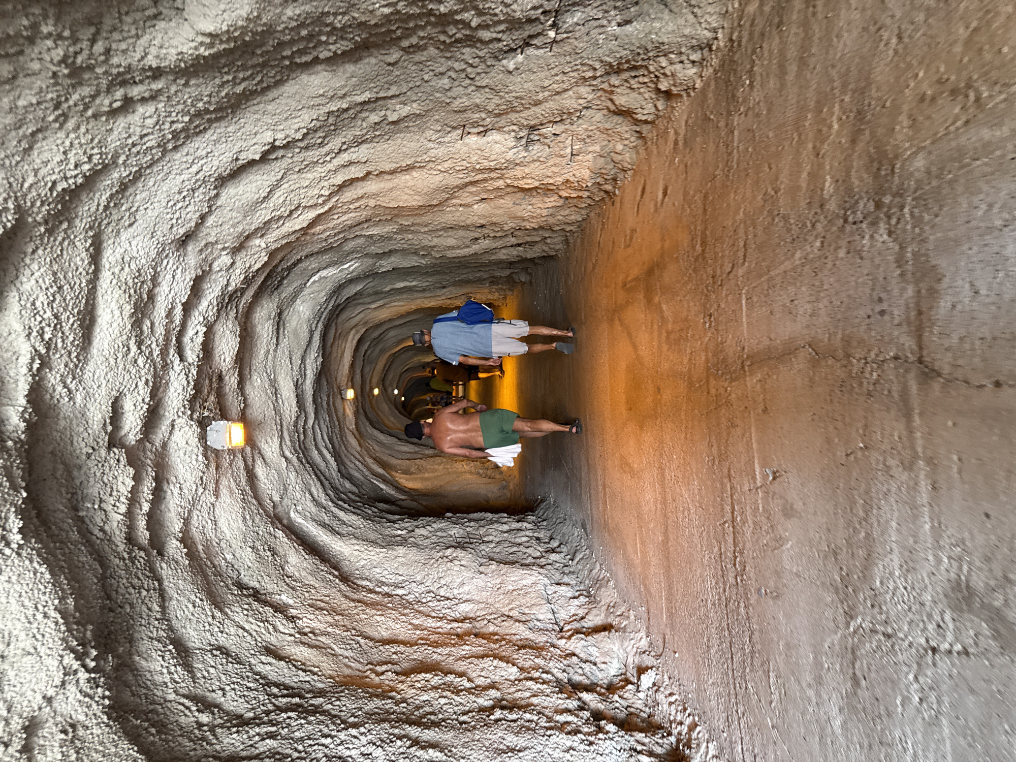The image depicts an interior view of a rough-hewn tunnel with a sandy floor. The tunnel walls are textured, appearing to be made of natural rock or a similar material, with visible ridges and undulations. Several lights are mounted on the ceiling, casting a warm glow that illuminates the path ahead.
In the foreground, two men are walking away from the camera. The man on the left is shirtless, wearing green shorts and carrying a white towel. The man on the right is wearing a blue t-shirt, light-colored shorts, and a blue bag slung over his shoulder. They appear to be walking in the same direction. Further down the tunnel, other figures can be seen, suggesting more people are present.
The tunnel appears to be a pedestrian walkway, possibly a natural cave passage or a man-made tunnel designed to resemble one. The presence of people in casual attire suggests a recreational or tourist setting. The overall impression is of a walk through a unique, natural or semi-natural environment.
The location is described as the city of Rafailovići, Montenegro. This specific tunnel could be a known pathway within the city's natural landscape or a man-made attraction. Without further context, it's difficult to pinpoint the exact location within Rafailovići. However, given the description and visual cues, it's likely a site accessible to visitors.