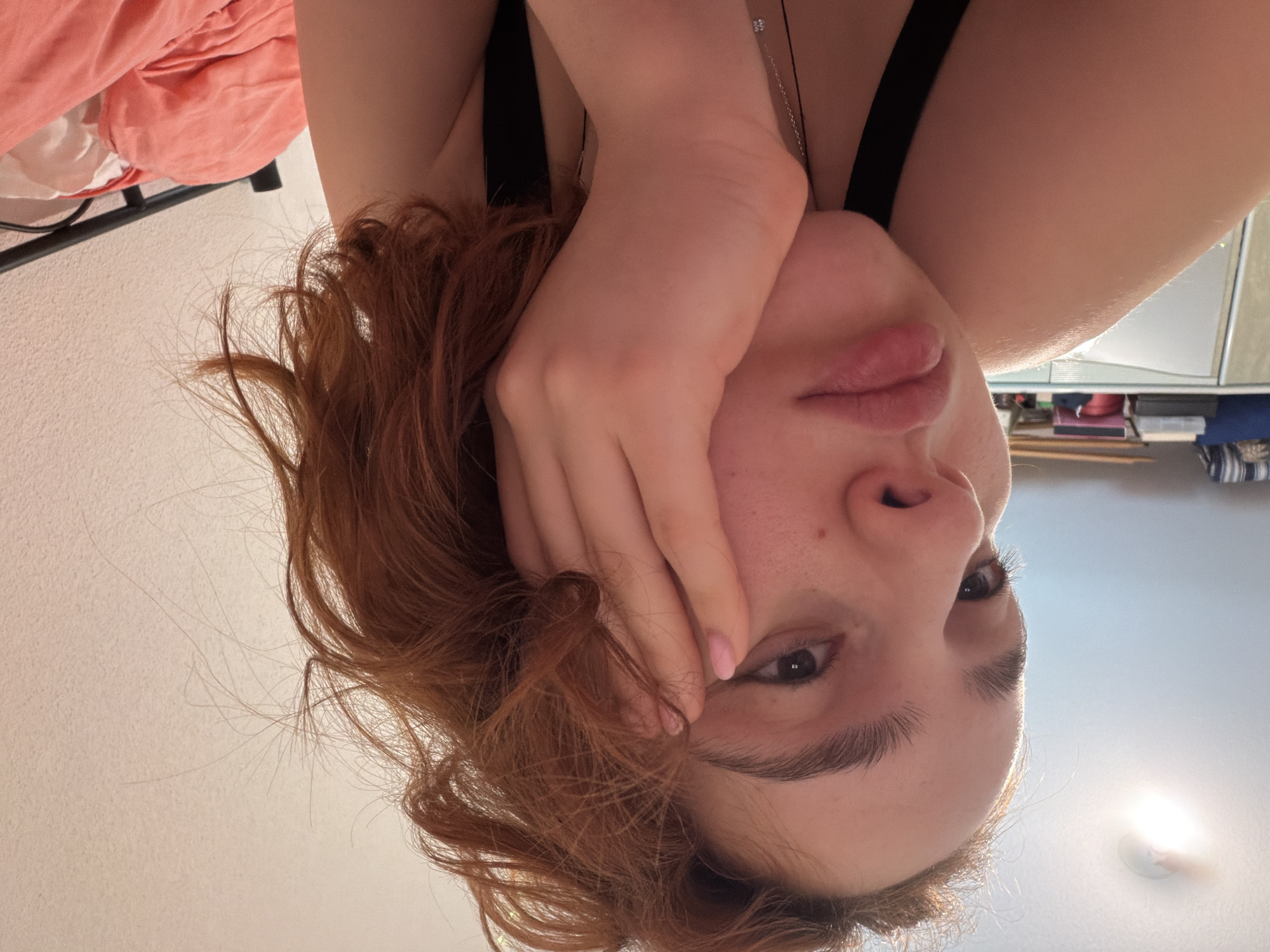 A close-up selfie shows a young person with auburn hair resting their face on their hand. They are wearing a black tank top and a silver necklace. The background consists of a pink wall, a bedside table with items on it, and a bed with pink bedding. The lighting appears to be from an overhead lamp. The scene is likely an indoor residential setting, possibly a bedroom. There is no indication of the location being in Strasbourg, France.