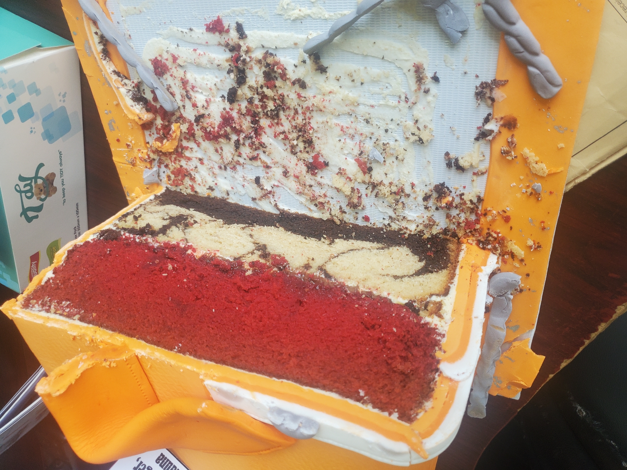 The image shows a close-up, slightly tilted view of a partially eaten cake. The cake is layered with red, vanilla, and chocolate cake, covered in orange and white frosting. Crumbs and frosting are scattered on a white paper liner and an orange paper base. The background is out of focus, showing a dark surface and a box with a teddy bear logo. There is no indication of the location within the city of Gwagwada, Nigeria.