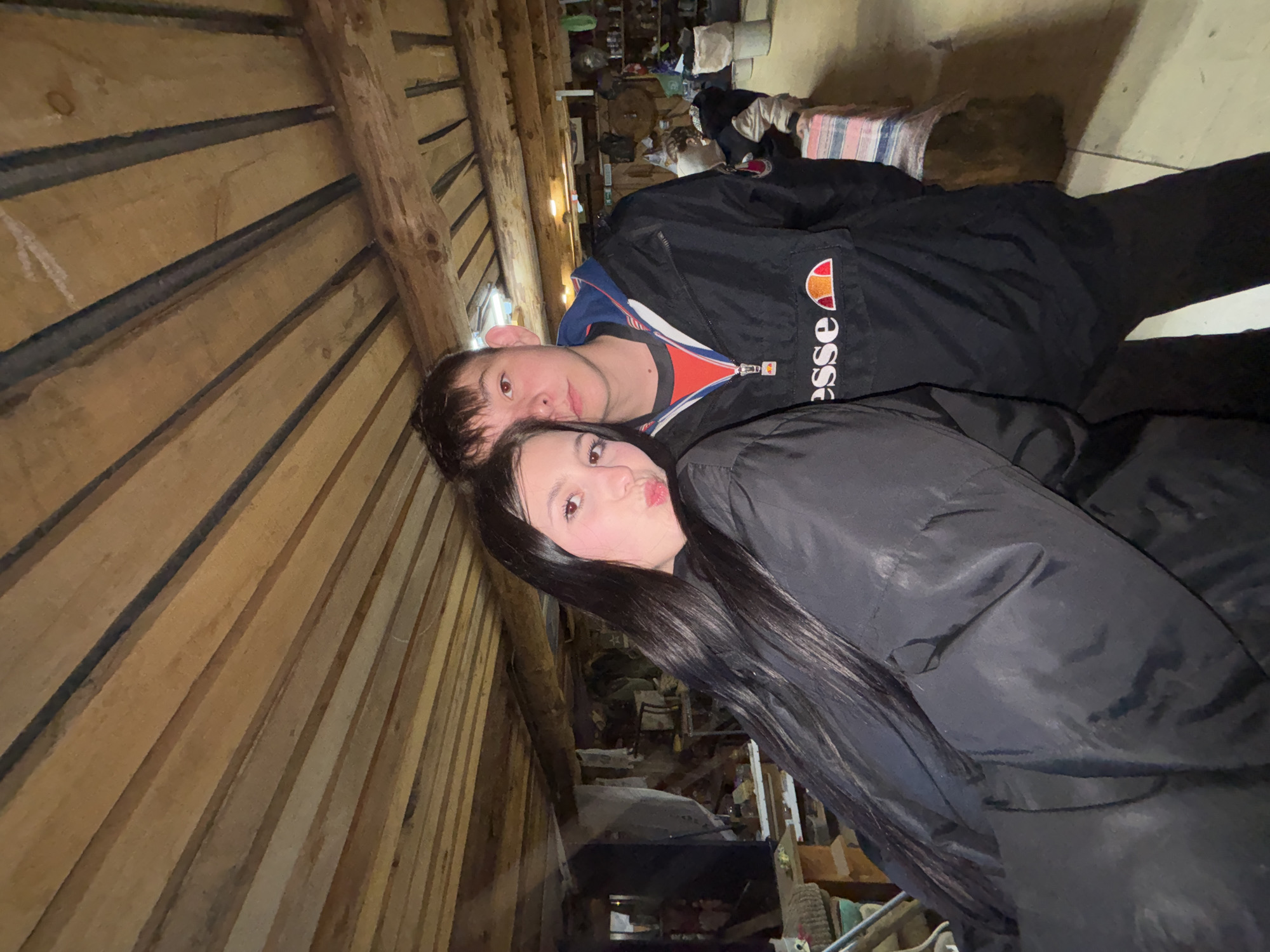 The image shows a selfie of two people, likely a couple, taken in what appears to be a cluttered, rustic interior. The wall behind them is constructed of wooden planks. There is a man with short dark hair wearing a jacket with an orange and white "Ellesse" logo. A woman with long dark hair and dark clothing is next to him, making a kissing face toward the camera. In the background, there are various objects and items stored or piled up, suggesting a storeroom or shed-like space. The location might be Фоки, Russia.