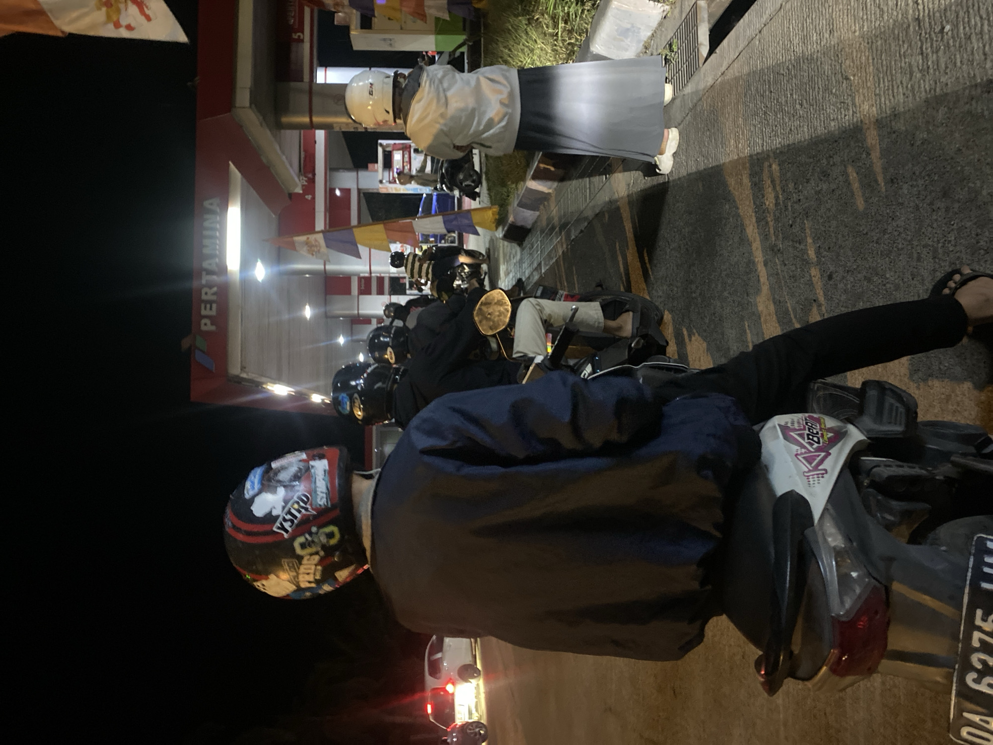 At night, in front of a Pertamina gas station in Gambut Barat, Indonesia, several people on motorcycles are lined up, some waiting to refuel. In the foreground, a person wearing a dark jacket and a helmet adorned with various stickers is seated on a white and black motorcycle. To the right, another individual in a white helmet and a long gray skirt stands beside a motorcycle. Behind them, more motorcyclists are visible, with the gas station's illuminated canopy overhead. The scene is illuminated by the gas station's lights and the headlights of nearby vehicles, creating a dimly lit, urban nighttime atmosphere.