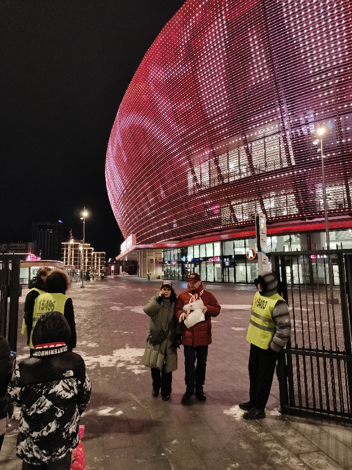 An outdoor night scene depicts people on a snow-dusted paved walkway in front of the illuminated Yekaterinburg Arena in Yekaterinburg, Russia. The stadium's large, curved facade dominates the right and background, glowing vibrant red with a grid pattern of white lights.
In the mid-ground, a couple walks toward the foreground-left. The woman wears a dark, long puffy winter coat, dark pants, and a dark hat, holding a phone to her right ear. The man next to her wears a red puffy winter jacket, dark pants, a red scarf, and a red baseball cap, carrying two white bags in his left arm.
To the far right, partially obscured by a dark metal gate, an individual in a yellow high-visibility vest with the number "15403" on the back stands facing partially away from the camera. Another dark metal gate is visible on the far left.
Further left, two additional individuals in yellow high-visibility vests are visible, one with the number "15496" clearly displayed on their back, standing near the left gate. In the immediate foreground, a person, likely a child, is seen from behind, wearing a black and white patterned winter coat and a dark hat with red lettering, holding a red object.
The ground is a light-colored paved surface, partially covered with patches of white snow or slush. Tall streetlights are illuminated. In the distant background, to the left, multiple modern buildings with illuminated windows and facades are visible against the dark night sky. The overall atmosphere suggests a post-event crowd departure or pre-event arrival at the stadium in cold weather.