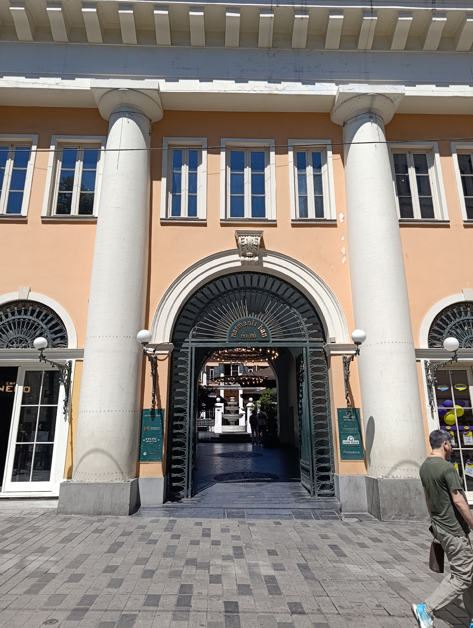 A man in a green t-shirt and khaki pants walks past a building with a large archway. The building has peach-colored walls, large white columns, and several windows. The archway is ornate with a black metal gate. There are signs on the building advertising businesses such as "Penguen Kitabevi," "Be Hunger," "Yildiz Kurye," and "Halkbank." The ground is paved with gray and black square tiles. The scene appears to be in Beyoğlu, Turkey.