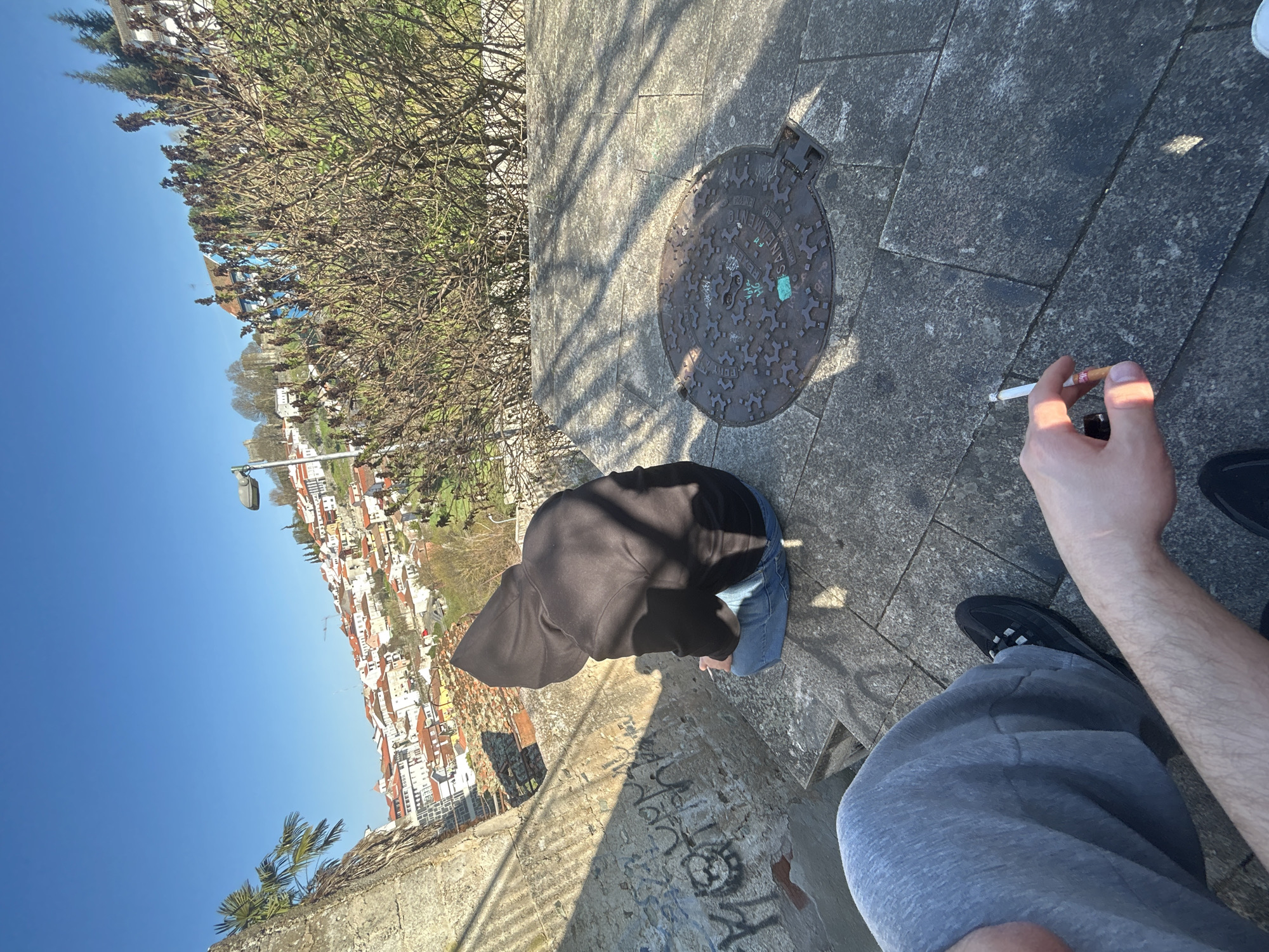 The image depicts an elevated outdoor urban setting in Bragança, Portugal, overlooking a city panorama under a clear blue sky.
In the immediate foreground, the lower torso and left arm of an individual are visible. This individual wears grey sweatpants and a black athletic shoe on their left foot. Their right hand holds a lit cigarette with an orange-brown filter.
In the midground, a second individual is seated on a stone surface, their back turned towards the camera. This person wears a black hooded sweatshirt and blue jeans. A small white object is visible near their right hand. To the left of the seated individual, a rough stone wall features visible graffiti in black and white. To their right, a circular cast-iron manhole cover with a patterned surface and the word "SANEAMENTO" is present on the stone ground. Strong diagonal shadows, indicative of direct sunlight, are cast across the stone surfaces, suggesting an overhead structure or fence.
The background presents a vast view of the city of Bragança, characterized by numerous buildings with predominantly red-tiled roofs sprawling across a hillside. Green foliage, including bare deciduous trees and some coniferous trees, is interspersed among the structures. A distant blue building is visible to the right. A streetlamp pole is present on the right side. On the far left, a palm frond is visible near a stone wall, likely part of the city's fortifications or an elevated platform. The sky is bright and clear blue.