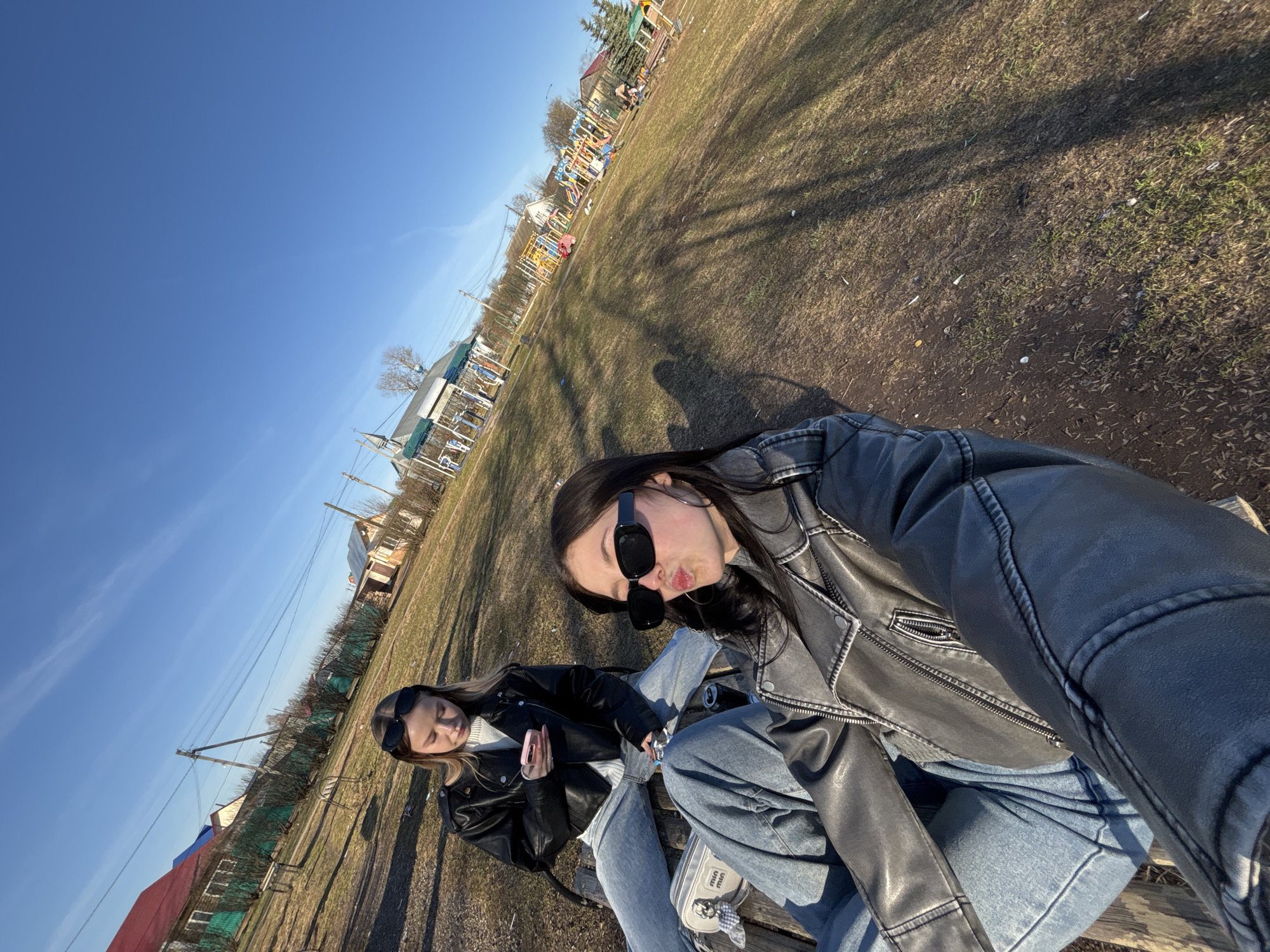 This image captures two young women sitting outdoors on a sunny day in what appears to be a park or open field in Фоки, Russia. The photo is taken from a high, tilted angle, likely a selfie.
The subjects are two young women. The one closer to the camera has long dark hair and wears black sunglasses, a black leather jacket, and blue jeans. She is making a playful expression with puckered lips and holds a silver drink can. A light-colored bag with "miu miu" text is visible next to her on the wooden bench. The second woman, with lighter long hair, is seated slightly further back, also wearing sunglasses, a black leather jacket, and blue jeans, looking down at a pink smartphone in her hands.
The setting is an outdoor area with sparse, dry grass and dirt, featuring long shadows that indicate either early morning or late afternoon. In the background, colorful playground equipment is visible, including slides and climbing frames. Beyond the playground, several houses, utility poles, and power lines can be seen against a clear blue sky. The overall scene suggests a relaxed, casual gathering on a bright day.