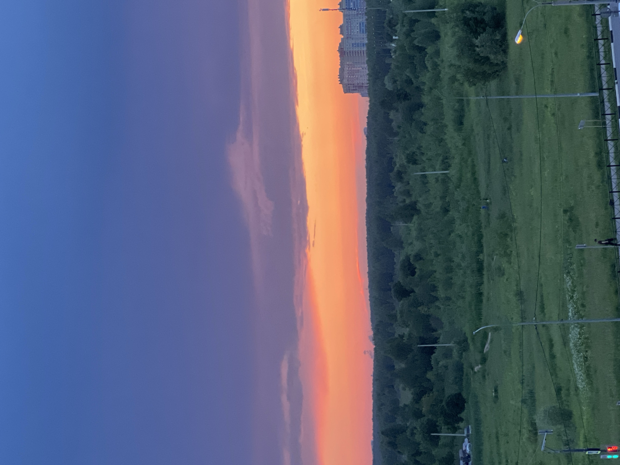 The image depicts an aerial view of a green landscape with a vibrant sunset in the background. The sky transitions from deep blue at the top to hues of purple and then a bright orange and pink band where the sun is setting. A dense forest occupies the middle ground, bordering a field of green grass. In the distance, a cluster of tall, multi-story buildings stands out against the horizon. The foreground shows a paved road with streetlights and a pedestrian walking. There are also traffic lights visible. The overall scene is natural with signs of urbanization.
