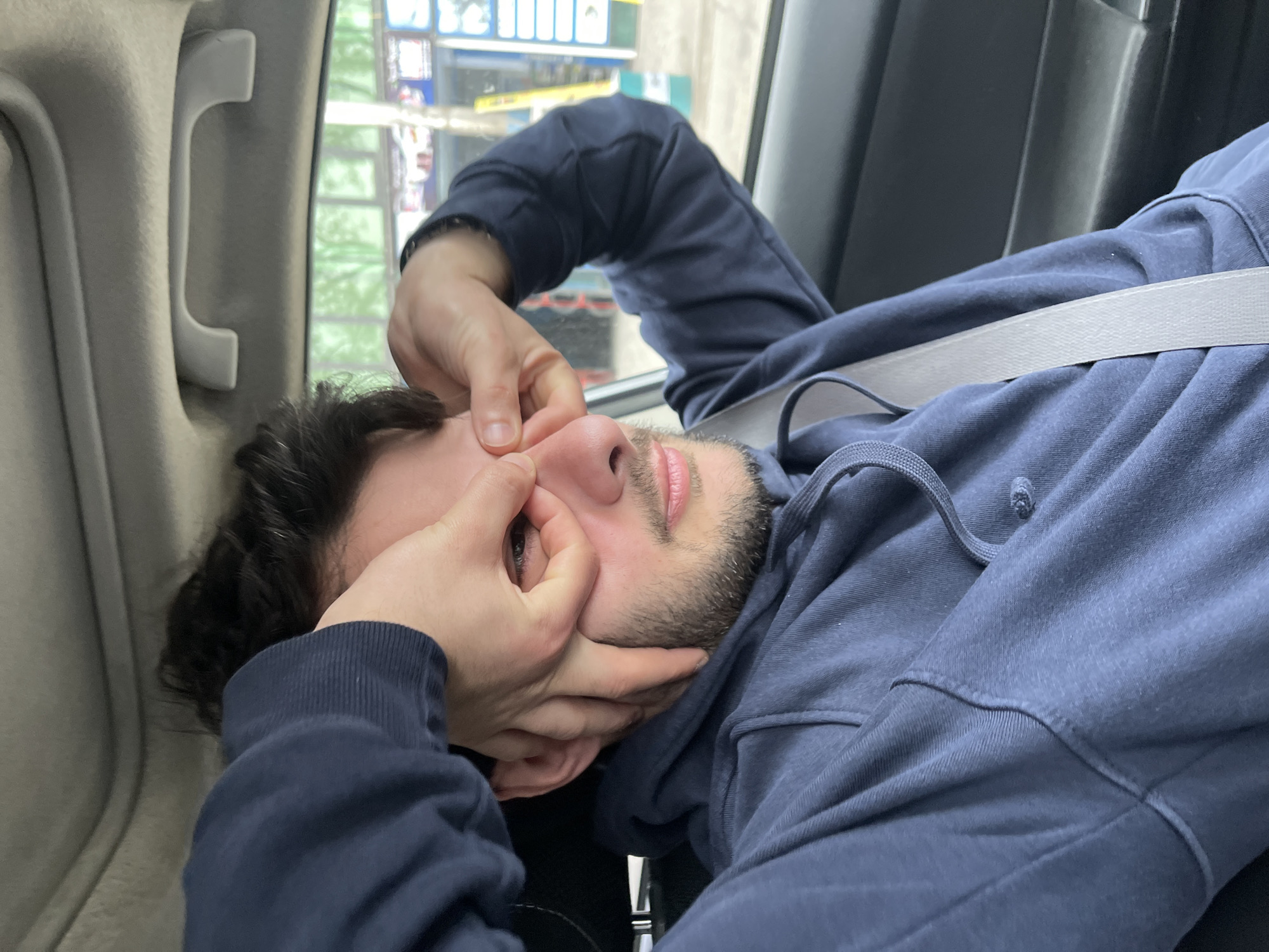 A man with dark curly hair and a beard is lying down in what appears to be a car, wearing a navy blue hoodie. His seatbelt is fastened. He is holding his face with both hands, fingers pinching the bridge of his nose and his cheeks. The location is suggested to be in Tbilisi, Georgia, based on the background viewed through the car window, which shows signs and building styles typical of the city. The car's interior is visible, including the overhead grab handle and a dark-colored seat behind him.