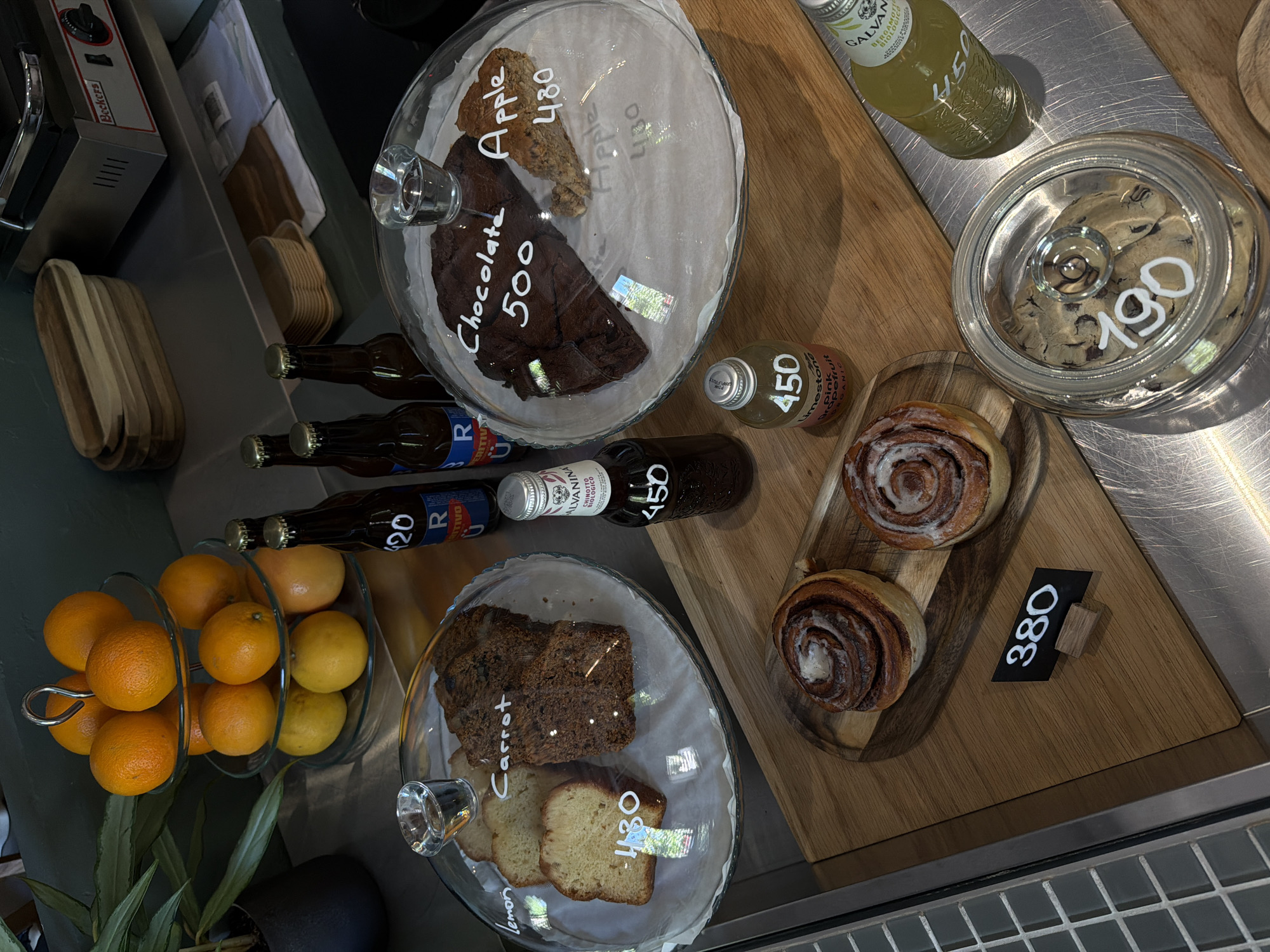 A top-down view shows a display of food items in a café setting. Oranges fill a tiered glass fruit holder. Brown bottles, labeled "R Eritivo 420" and "Alvanina Chinotto 450," lie horizontally next to a glass dome containing carrot and lemon bread. Underneath the glass dome, handwritten labels indicate "Carrot" and "lemon." Two cinnamon rolls lie on a wooden board with a black price tag indicating "380." A glass jar with cookies is nearby, marked with a price of "190". Bottles of "Galvanina Bergamotto Organico 450" and "Limestone Pink Grapefruit Organic" are positioned next to another dome filled with "Chocolate" and "Apple" pieces, priced at "500" and "480," respectively.
There is no mention or visual evidence to suggest that the cafe is located in the city of Belgrade, Serbia.