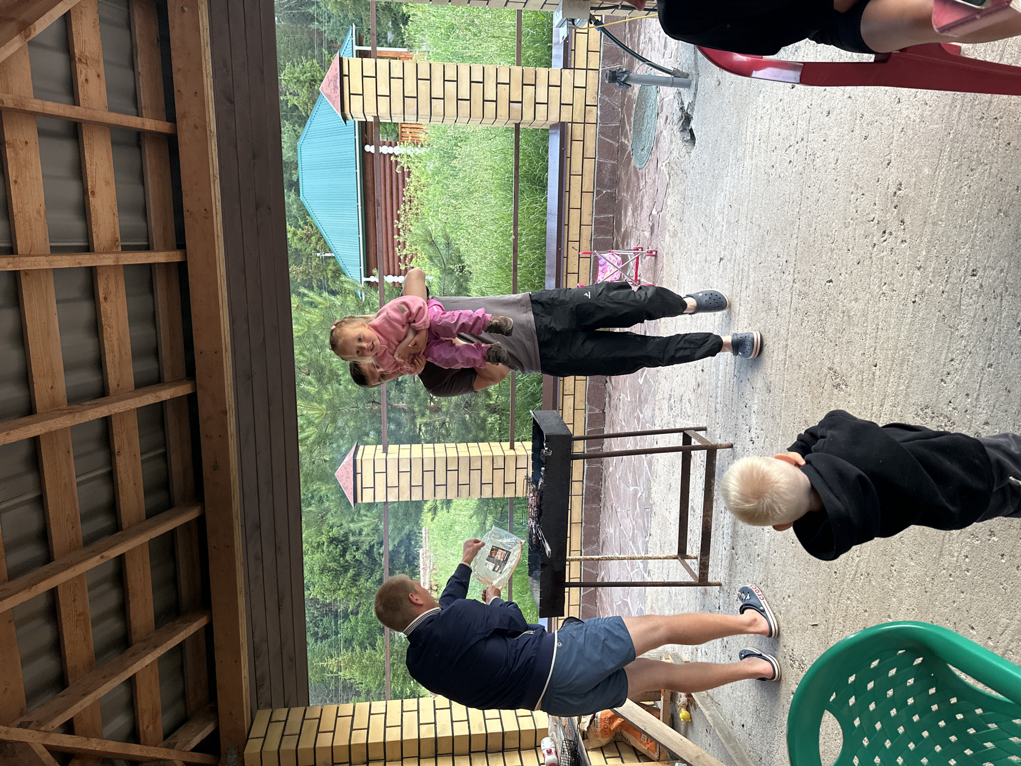 In the city of Pepelyaeva, Russia, a man in a dark blue jacket and blue shorts is standing by a black barbecue grill, holding a clear plastic bag and appearing to be adding something to the grill. He is wearing blue Croc-style sandals. To his right, another man in a grey t-shirt and black pants is holding a young child wearing a pink sweater and pink pants. The child is looking towards the camera. In the foreground, a young boy with blond hair, wearing a black hoodie, is facing away from the camera, with his hands to his ears. The scene takes place outdoors under a wooden roof structure with a metal roof. Behind the barbecue, there is a brick wall and a natural landscape with trees and grass, and a building with a teal roof. There are also several chairs visible, including a green plastic chair in the lower left and a red chair on the right. The ground is paved with concrete.