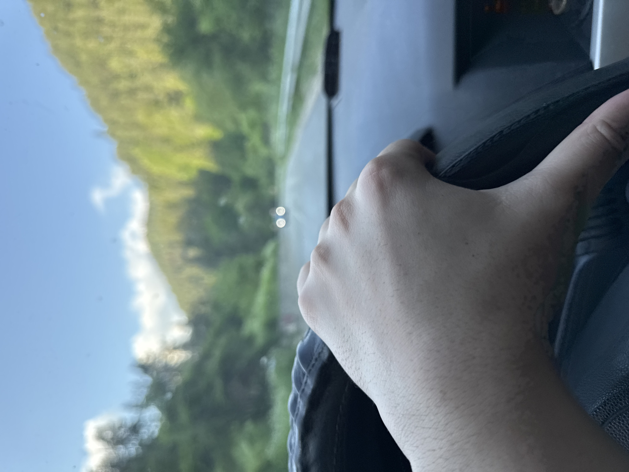 The image captures a first-person perspective from inside a vehicle, looking forward through the windshield into a natural landscape in Sokolovtsi, Bulgaria. In the immediate foreground, a person's light-skinned right hand, with visible hair on the knuckles and forearm, firmly grips a black, textured steering wheel. The visible portion of the arm exhibits irregular pigmentation or freckles. Part of the dark vehicle dashboard is visible below the hand.

The midground reveals a paved road, which appears slightly reflective, extending into the distance. Two distinct, circular, blurry white lights, indicative of an approaching vehicle's headlights, are visible directly ahead on the road. A light-colored guardrail runs along the right side of the road. The background is significantly out of focus, showing lush green, tree-covered mountains or hills flanking both sides of the road. Above the hills, the sky is clear blue with scattered white clouds. The overall scene depicts driving through a scenic, forested area.