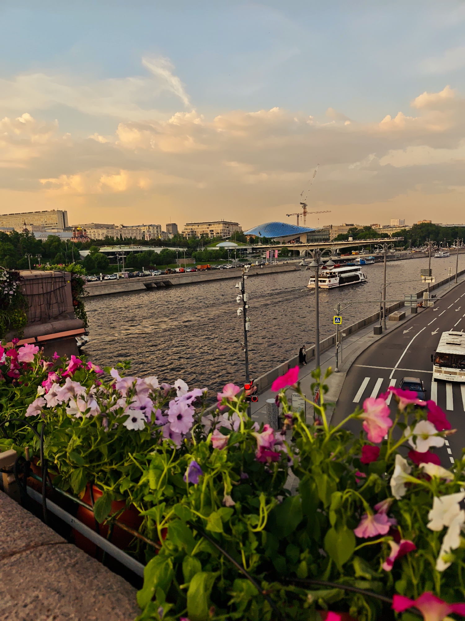 The image shows a cityscape at dusk, viewed from a high vantage point. In the foreground, a flower box filled with pink, purple, and white petunias decorates a stone wall. Beyond the wall, a wide river flows through the city, with a large passenger boat moving along its surface. On the right side of the river, a road runs parallel, with cars and a bus visible. Pedestrian crosswalks are marked on the road. Buildings of varying heights and architectural styles line the riverbanks, with a blue-domed structure and a crane in the distance. The sky is filled with clouds tinged with orange hues from the setting sun.
