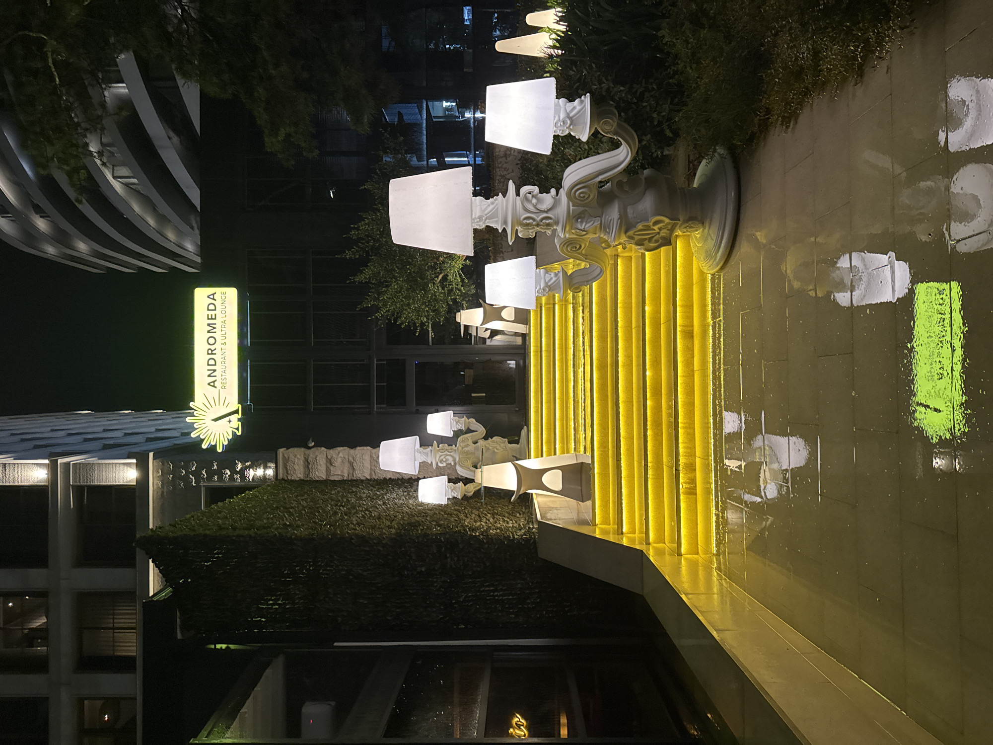 The image depicts an outdoor scene at night, likely in Budva, Montenegro, based on the "Andromeda Restaurant & Ultra Lounge" sign visible on the left. The area appears to be wet, possibly from rain, as evidenced by the reflections on the ground. A row of ornate, illuminated white lamps lines a set of yellow-lit stairs leading upwards. Vegetation is present on both sides of the stairs, with a building visible in the background.