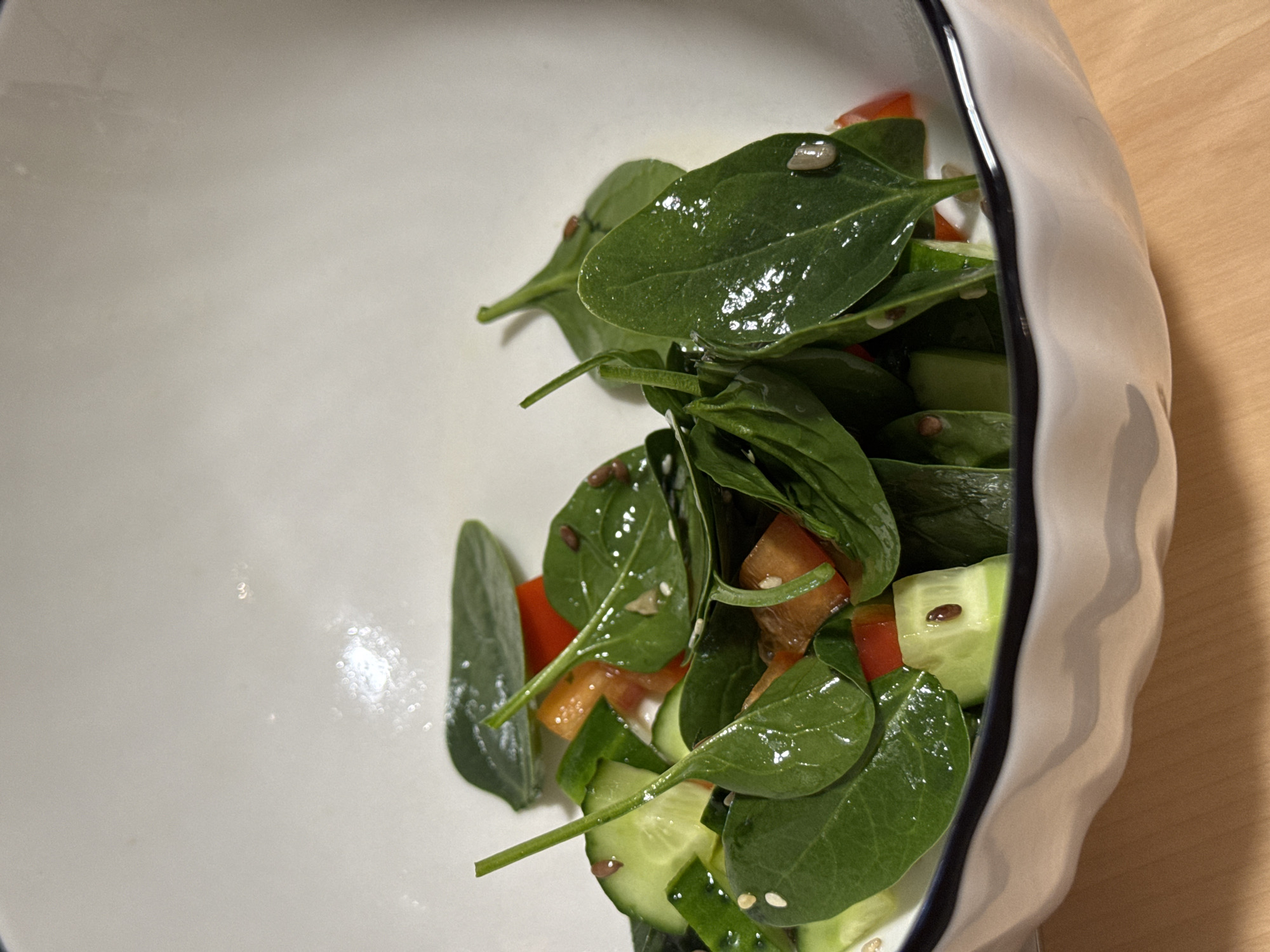 A salad consisting of spinach, cucumber, and tomato is in a white bowl. The salad is dressed with oil and sprinkled with seeds. The bowl is sitting on a wooden surface. The location of the city of Izhevsk, Russia is not discernible from the image.