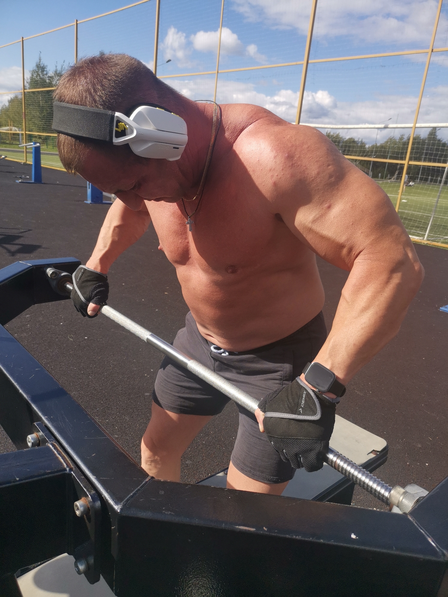 The image shows a muscular shirtless man wearing a black headband, white headphones, and black weightlifting gloves. He is holding a barbell, appearing to be in the middle of an exercise. He is wearing black shorts and a silver watch on his left wrist. The man is positioned at an outdoor gym facility with a black metal apparatus in the foreground. In the background, there is a fence, trees, a sports field, and a blue sky with clouds. The scene is set in the city of Kizel, Russia, indicated by information not present in the image itself.