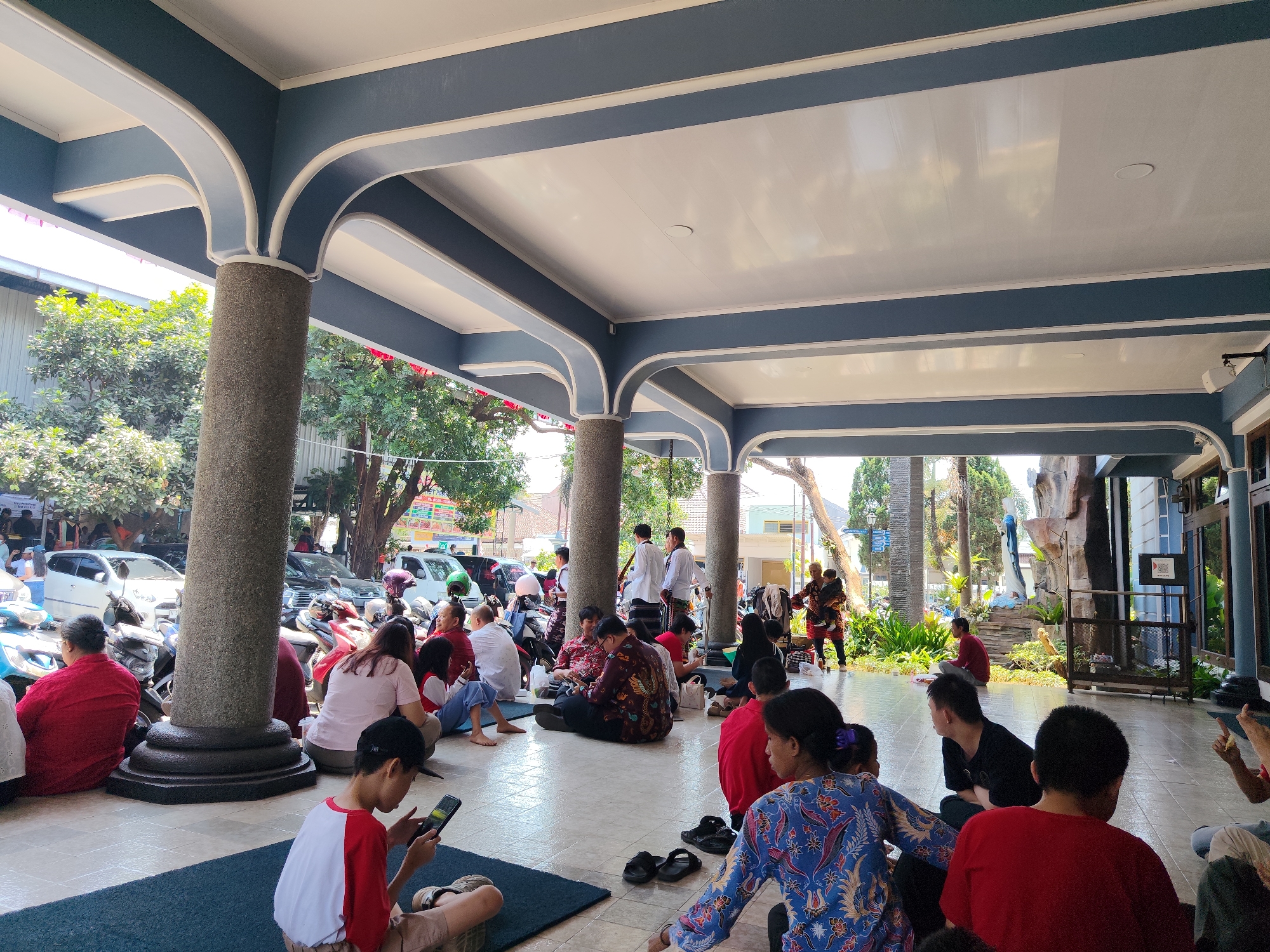 The image depicts a gathering of people seated on the floor under a covered outdoor area with a white ceiling and blue trim. Several large, textured stone pillars support the roof. The ground is tiled. In the background, there are trees, parked cars, and motorcycles. Some individuals are interacting with their phones. The scene appears to be in a public space, possibly a religious site or a community gathering area. The presence of specific architectural elements and the general ambiance suggest it could be located in Malang, Indonesia.