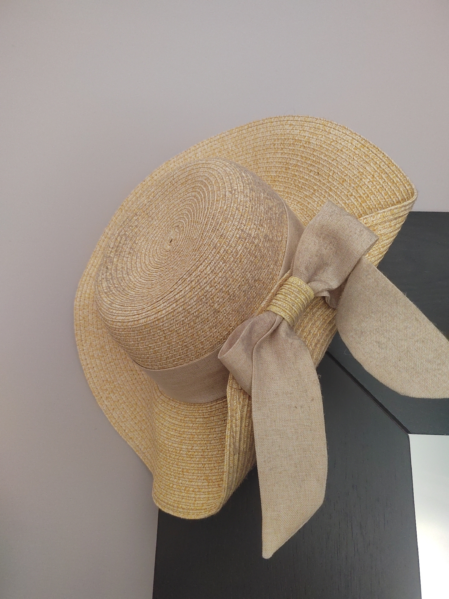 This media file features a straw hat with a beige ribbon tied in a bow. The hat is positioned on a dark surface, likely a piece of furniture, and a pale wall is visible in the background. The hat is angled, with its brim extending downwards and to the right. The ribbon has a looped bow with two trailing ends, one of which is more prominent. The texture of the straw and the ribbon is visible. There are no people or animals present, and no specific activity is taking place. The lighting appears to be indoor ambient light, without any strong shadows. The setting is nondescript, offering no specific clues about the location other than what is provided in the prompt. There is no visible text in the image.