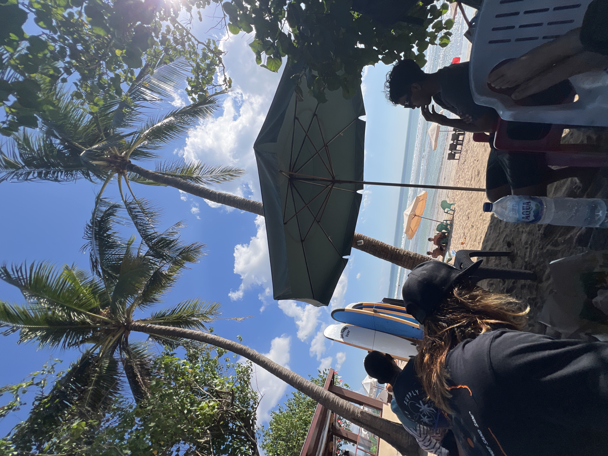 The image depicts a sunny day at a beach in Kuta, Indonesia. In the foreground, three individuals are seated on chairs under the shade of a large green umbrella, facing away from the camera. One person, in a black t-shirt with visible text, has blonde hair and is wearing a black cap. Next to them, another person in a dark t-shirt with a graphic design appears to be looking out towards the ocean. A third person, seated on the right, is wearing sunglasses and a dark t-shirt, resting their chin on their hand. A bottle of Aqua water is placed on the sand near the seated individuals.
Behind them, two surfboards stand upright, leaning against each other. Further down the beach, several beach umbrellas are set up, with a few people relaxing on chairs or walking near the water. The ocean is visible in the background with small waves. Palm trees are scattered throughout the scene, with some foliage creating a canopy over the foreground area. A building structure with a thatched roof is visible on the left side of the image. The sky is clear blue with scattered white clouds.