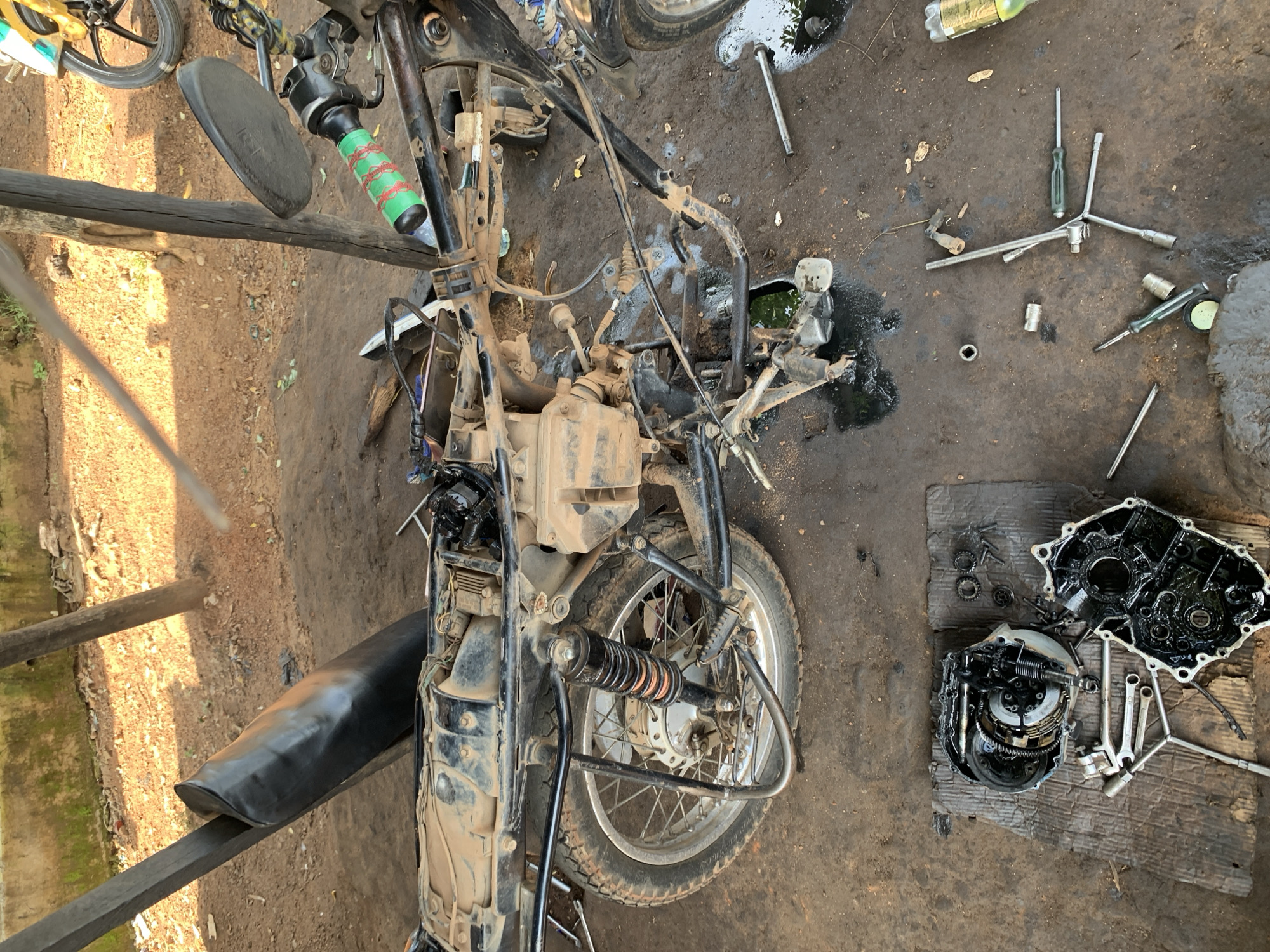 The image shows a partially disassembled motorcycle on a dirt ground, likely in a workshop or outdoor repair area. The motorcycle is dirty and appears to be undergoing maintenance. Several tools, including screwdrivers and wrenches, are scattered on the ground around the vehicle. A bottle and a plastic container with green liquid are also visible. The scene suggests a manual repair process, with components of the engine, specifically the clutch assembly, laid out separately. The lighting indicates it is daytime. There is no visible text in the image.