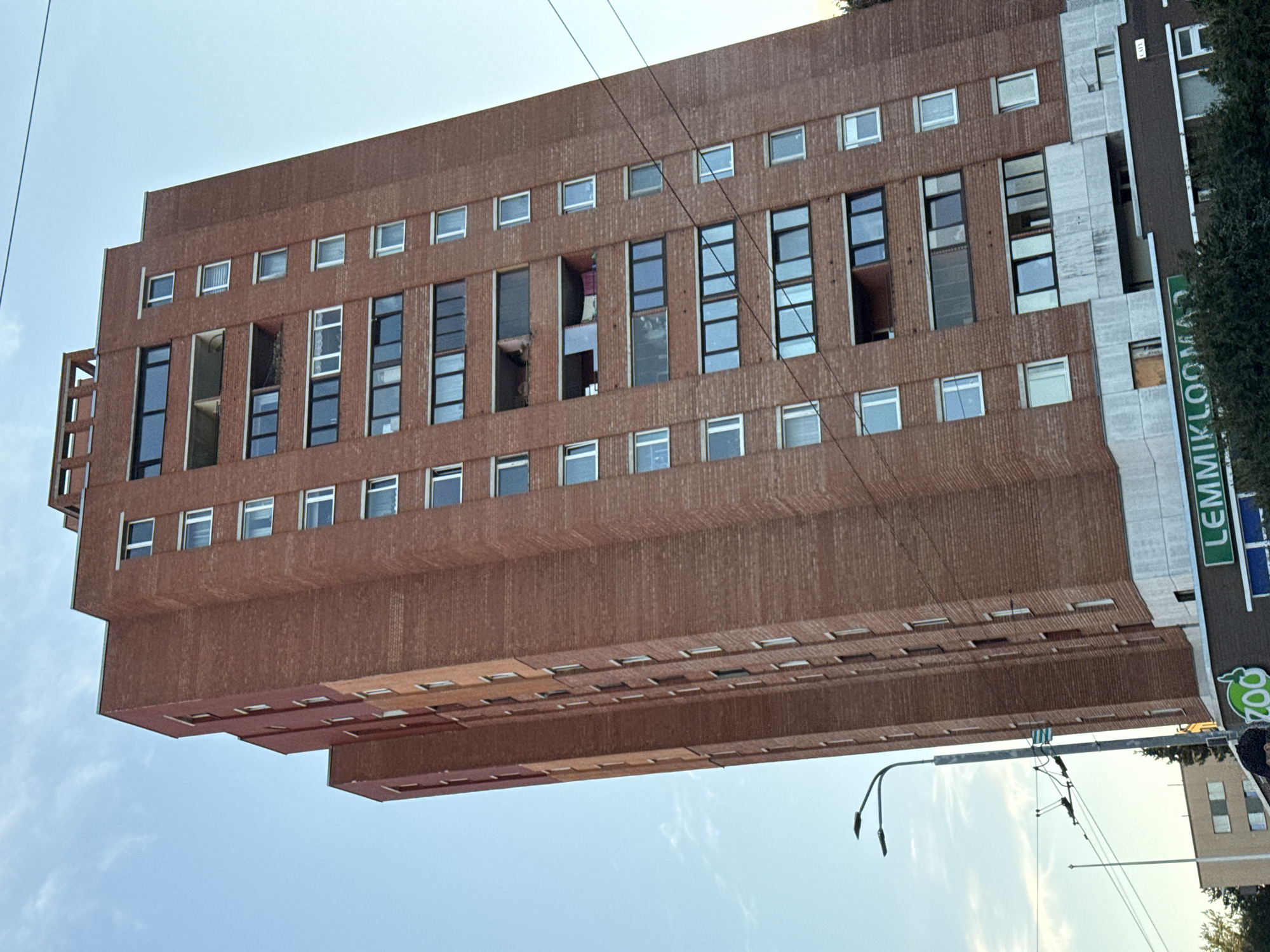 An eye-level, medium shot captures a tall, brick apartment building in Tallinn, Estonia, under a clear, blue sky with scattered clouds. The building is predominantly red brick and features numerous windows of various sizes and configurations, some with balconies. The perspective is slightly tilted, emphasizing the height of the structure. To the left, a street lamp with two lights and tram power lines are visible. At the base of the building, a storefront with green signage reads "LEMMIKLOOMA" and a green "ZO0" logo is present. In the background, another building and trees can be seen. The lighting suggests it is daytime, possibly late afternoon or early evening.