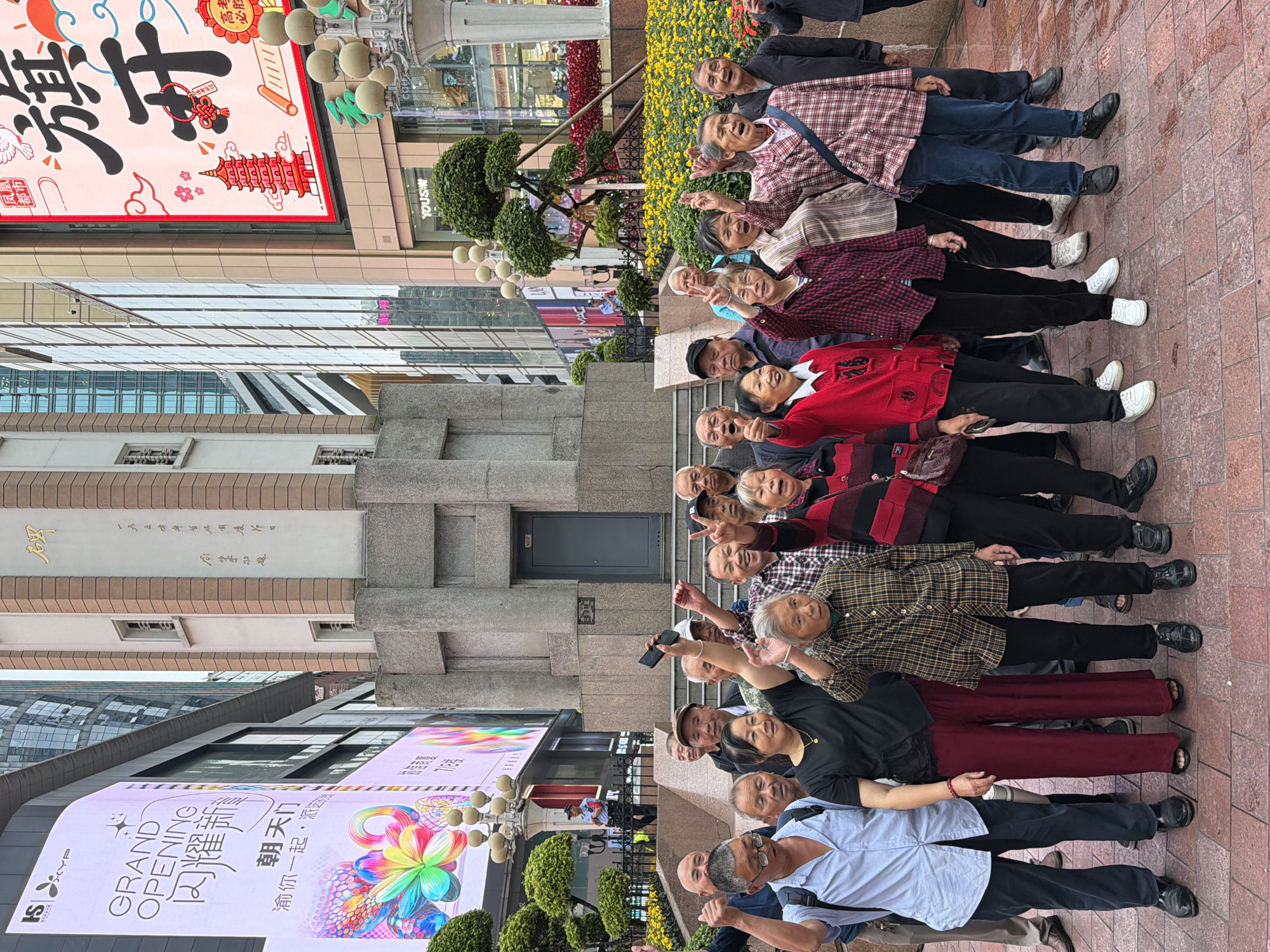 A group of approximately 20 elderly individuals, consisting of both men and women, are gathered outdoors in what appears to be a public square or pedestrian area. They are posing for a photograph, with many of them raising their hands and smiling. Most individuals are dressed in casual or traditional Chinese attire.
The background features modern city architecture, including tall buildings with glass facades and large digital billboards displaying colorful graphics and text in Chinese. One billboard prominently advertises a "GRAND OPENING." A cylindrical structure with ornate detailing, possibly a monument or clock tower, is also visible, along with streetlights and lush yellow and red flowers planted in garden beds. The ground is paved with brick-like tiles. A police officer is visible in the distance, standing near a barricade. The scene suggests a lively urban environment in Yuzhong District, China.