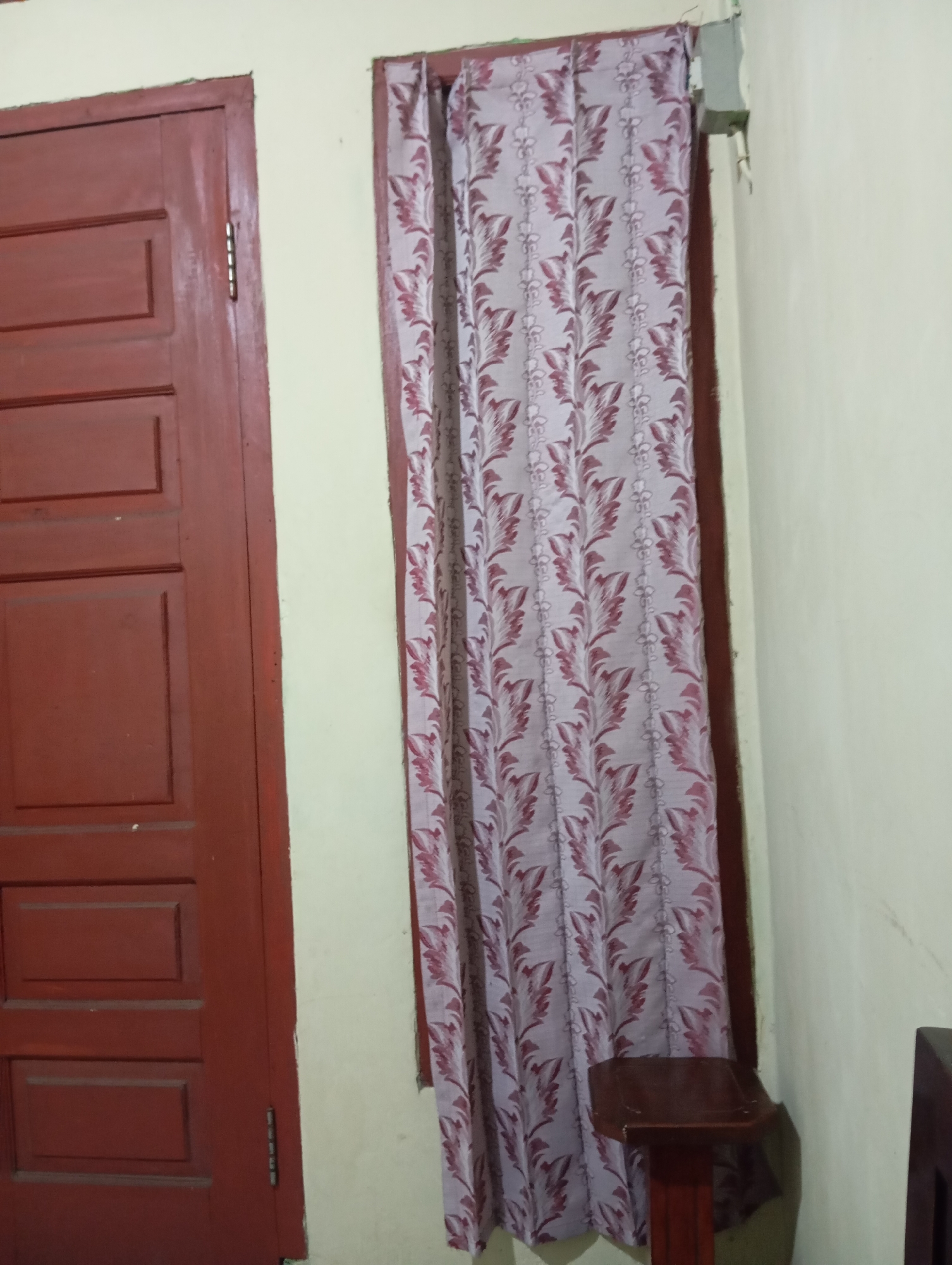 The image depicts an interior scene, likely a room in a house, in Bokol, Indonesia. On the left side of the image, a closed, dark red wooden door is visible. The door has a paneled design with three distinct horizontal rectangular sections. The hinge of the door is visible on the left edge of the frame. To the right of the door, a window is partially covered by a light-colored curtain with a repeating pattern of maroon-colored leaves and small floral motifs. The curtain is drawn partially closed, revealing a sliver of the window frame. The wall to the right of the curtain is painted a light, neutral color, possibly cream or off-white. At the bottom right of the image, a dark wooden stool or small table with a decorative edge is partially visible. The lighting in the scene appears to be artificial and somewhat dim, casting soft shadows. The overall impression is one of a modest, lived-in space.