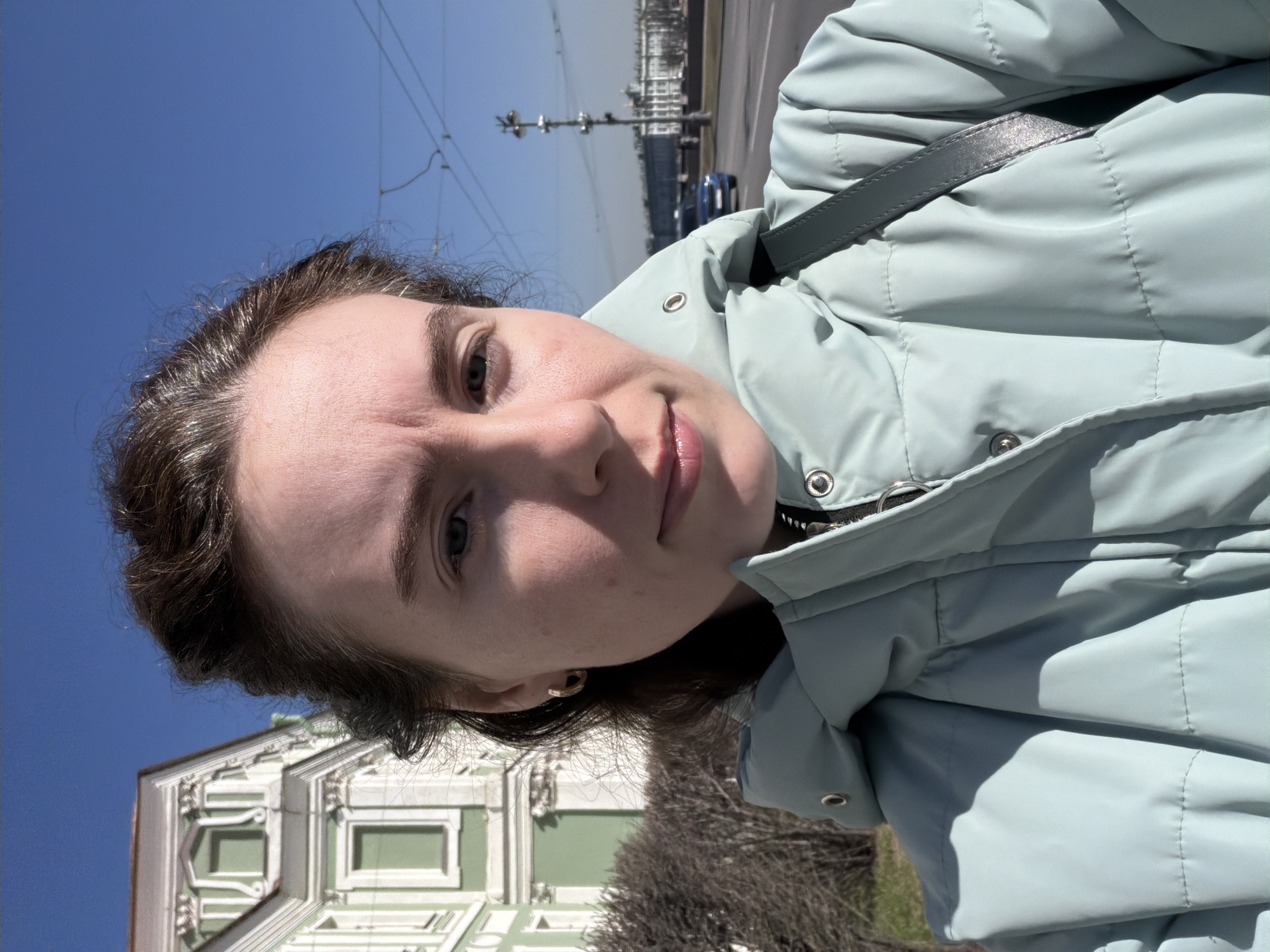 A woman takes a selfie. She is wearing a light blue quilted jacket and a gray purse or bag strap. The background shows a clear blue sky, a street with vehicles, and the Winter Palace in Saint Petersburg, Russia, which is identifiable by its green and white facade and architectural details. Some bare trees are visible. The photograph is rotated counterclockwise.