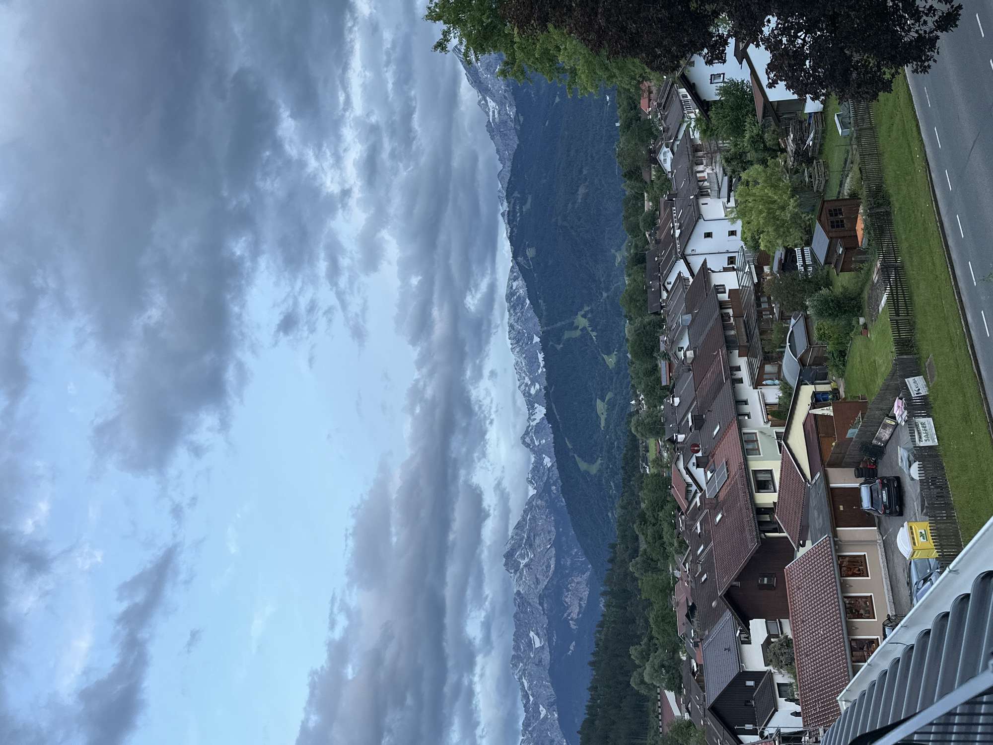 The image depicts a residential area in Garmisch-Partenkirchen, Germany, with a backdrop of snow-capped mountains. In the foreground, rooftops of houses are visible, many with brown or dark gray tiles. Some houses have visible solar panels. A gray paved road runs along the bottom right of the image, with white dashed lines indicating lanes. Adjacent to the road are green lawns and wooden fences. A dark-colored SUV is parked in a driveway, along with a yellow portable toilet and other assorted items. Several small wooden sheds are scattered throughout the yards. In the mid-ground, a dense collection of houses forms a neighborhood. Beyond the houses, rolling green hills are covered in trees, and further in the distance, the majestic Zugspitze massif mountains rise, their peaks dusted with snow. The sky is overcast with dramatic, dark gray clouds, suggesting an impending storm or the aftermath of one. The overall scene conveys a sense of a tranquil alpine town under a moody sky. There are no people visible in the image.
