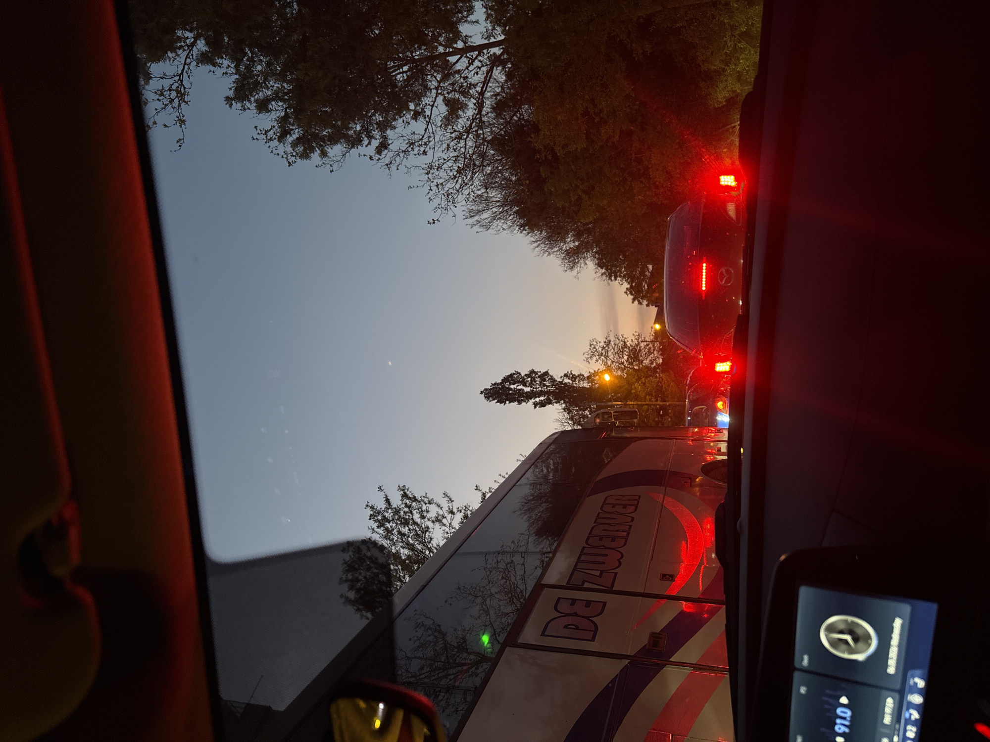 The image depicts a scene from inside a car, looking out through the windshield. The car is in traffic, with a bus to the left and a car directly ahead, visible through the windshield. The bus has the name "DE ZWERVER" on its side. It appears to be dusk or twilight, with trees visible above the vehicles. The car's dashboard and infotainment system are visible in the foreground. The location is Unknown, Unknown.