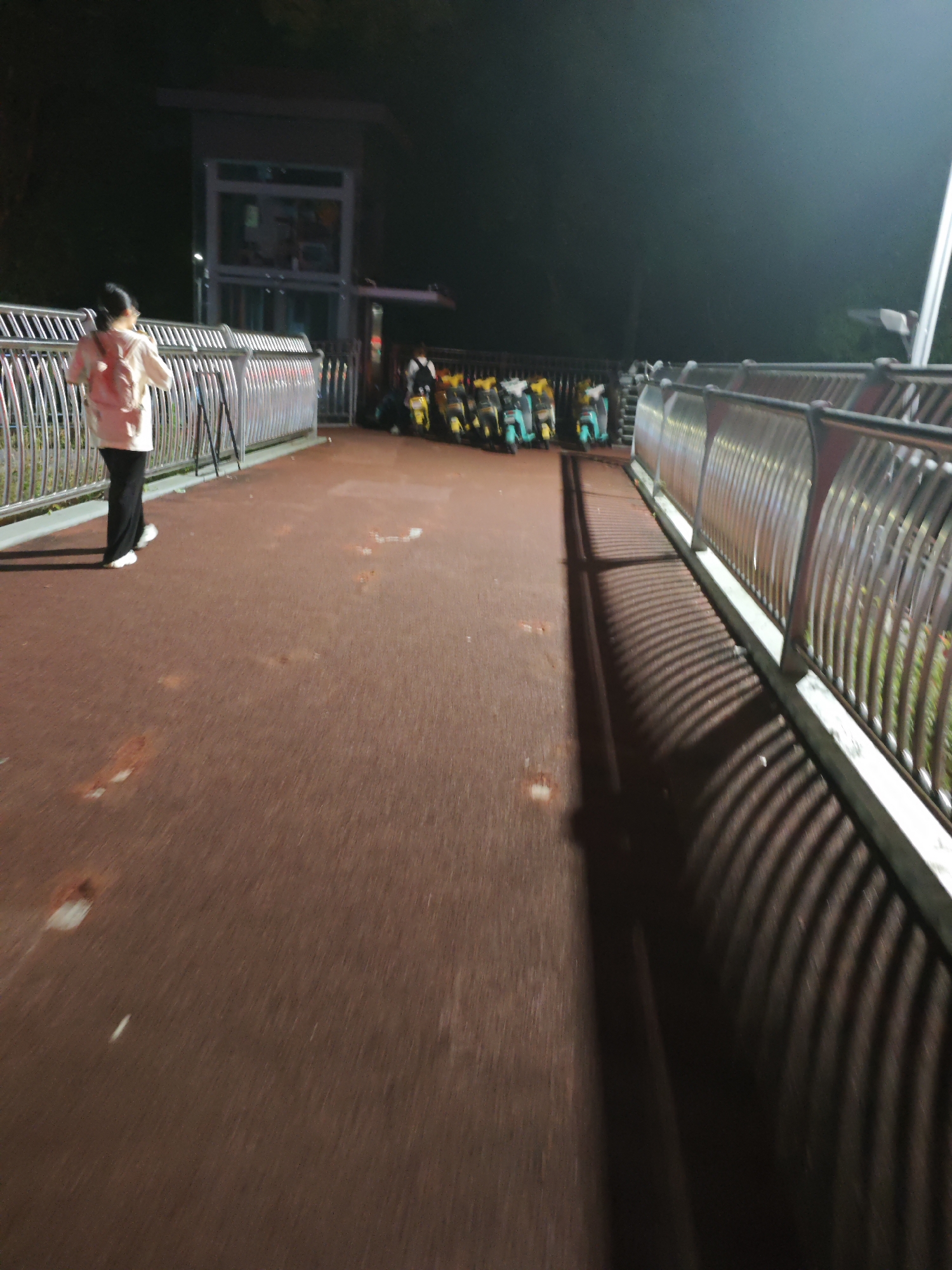 The image shows a person wearing a pink backpack and a white shirt, walking on a reddish-brown pathway at night. The pathway appears to be an elevated walkway or bridge, with metal railings on the sides. In the background, there are several electric scooters parked in a row. A tall structure, possibly a bus stop or a small building with glass windows, is visible further in the background. The scene is dimly lit, with artificial lighting casting shadows on the pathway. The location is identified as the Chongqing High-tech Zone, China.