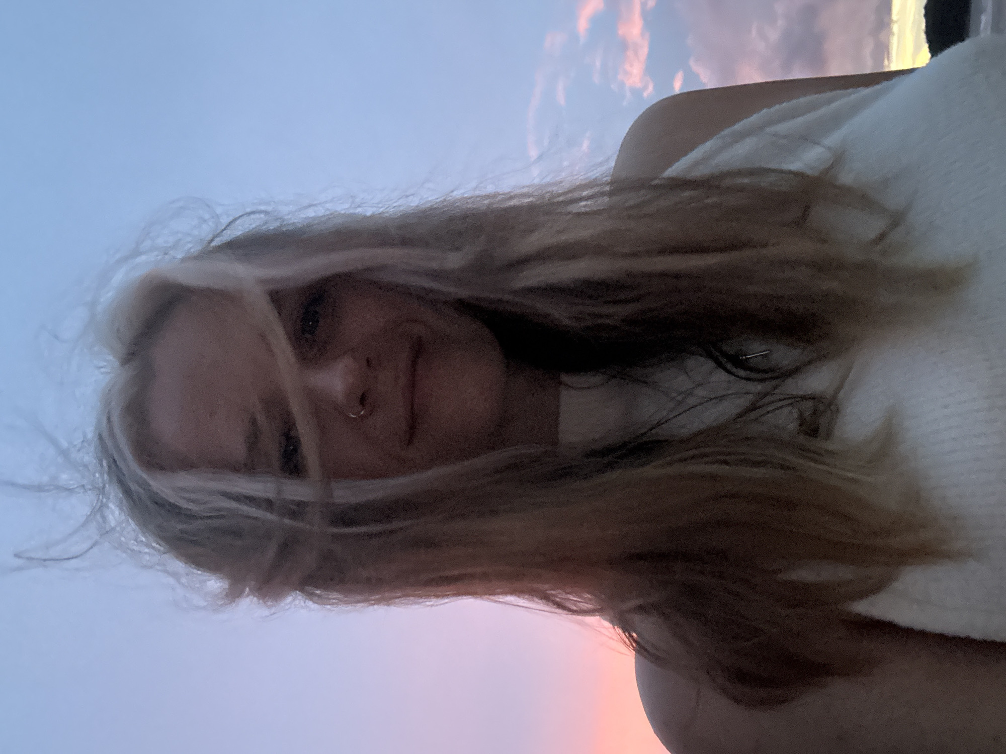 A young woman with blonde hair and a nose ring smiles at the camera. She is wearing a white top. Her hair is being blown by the wind. The background is a pastel sky with clouds, suggesting a sunset or sunrise. The location appears to be a beach or coastal area, possibly in Choeng Thale, Thailand, given the presence of a distant landmass on the right side of the frame.