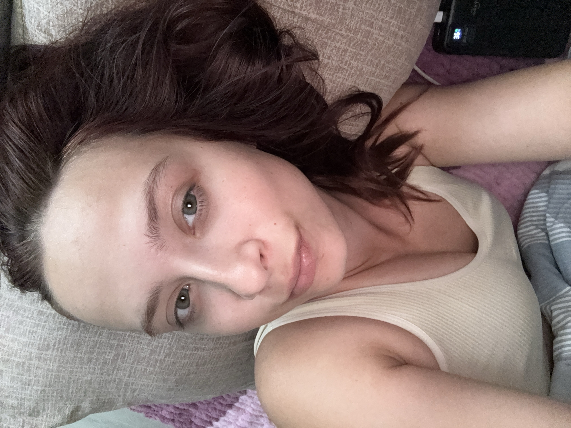 The image presents a relaxed, overhead view of a woman lying down indoors, likely in a bedroom setting. She has shoulder-length, wavy dark brown hair that spreads across a light grey or beige textured pillow. Her fair skin, light green eyes, and natural-looking eyebrows are prominent as she looks directly at the camera with a neutral, calm expression. Her lips appear softly moisturized.
She is wearing a simple, light cream-colored ribbed tank top. The background features various soft furnishings: a soft purple textured blanket beneath her and a blue and white striped blanket partially covering her on the right. In the upper right corner, a black electronic device, appearing to be a power bank, is visible. Its small screen displays "38%" and a "°C" symbol, indicating battery percentage and possibly a temperature reading.
The lighting is soft and ambient, suggesting an indoor environment, potentially during the morning or evening. The overall scene conveys a sense of comfort and repose. The location context provided is Chaikovsky, Russia.