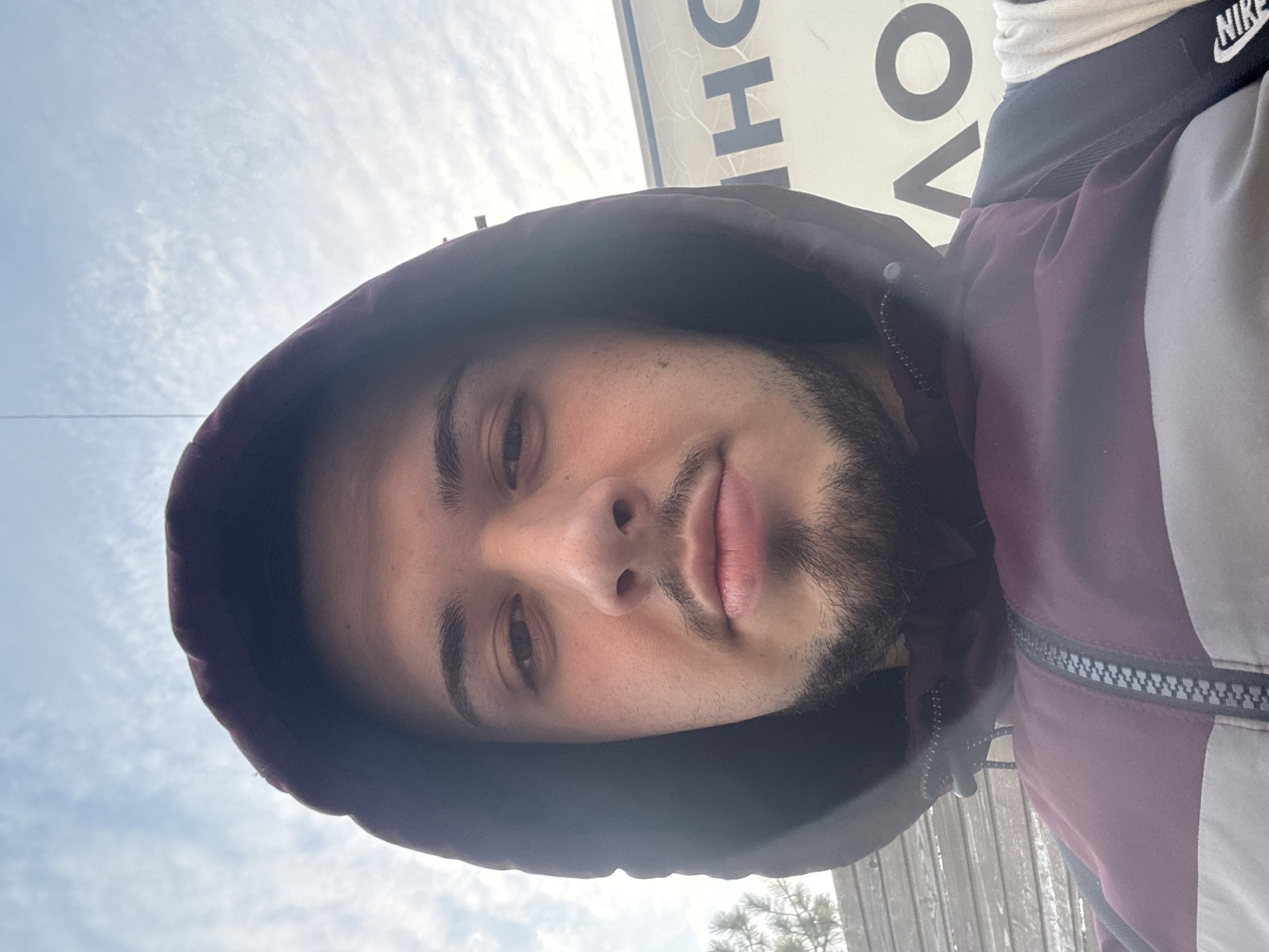 A selfie taken by a young man with medium skin tone, dark brown eyes, and a short, dark beard. He is wearing a maroon and gray hooded jacket with the hood up. The background includes a cloudy sky and a sign with large, partially visible letters. The Nike logo is visible on the back of his neck. The location in Sofia, Bulgaria is not discernible from the image.