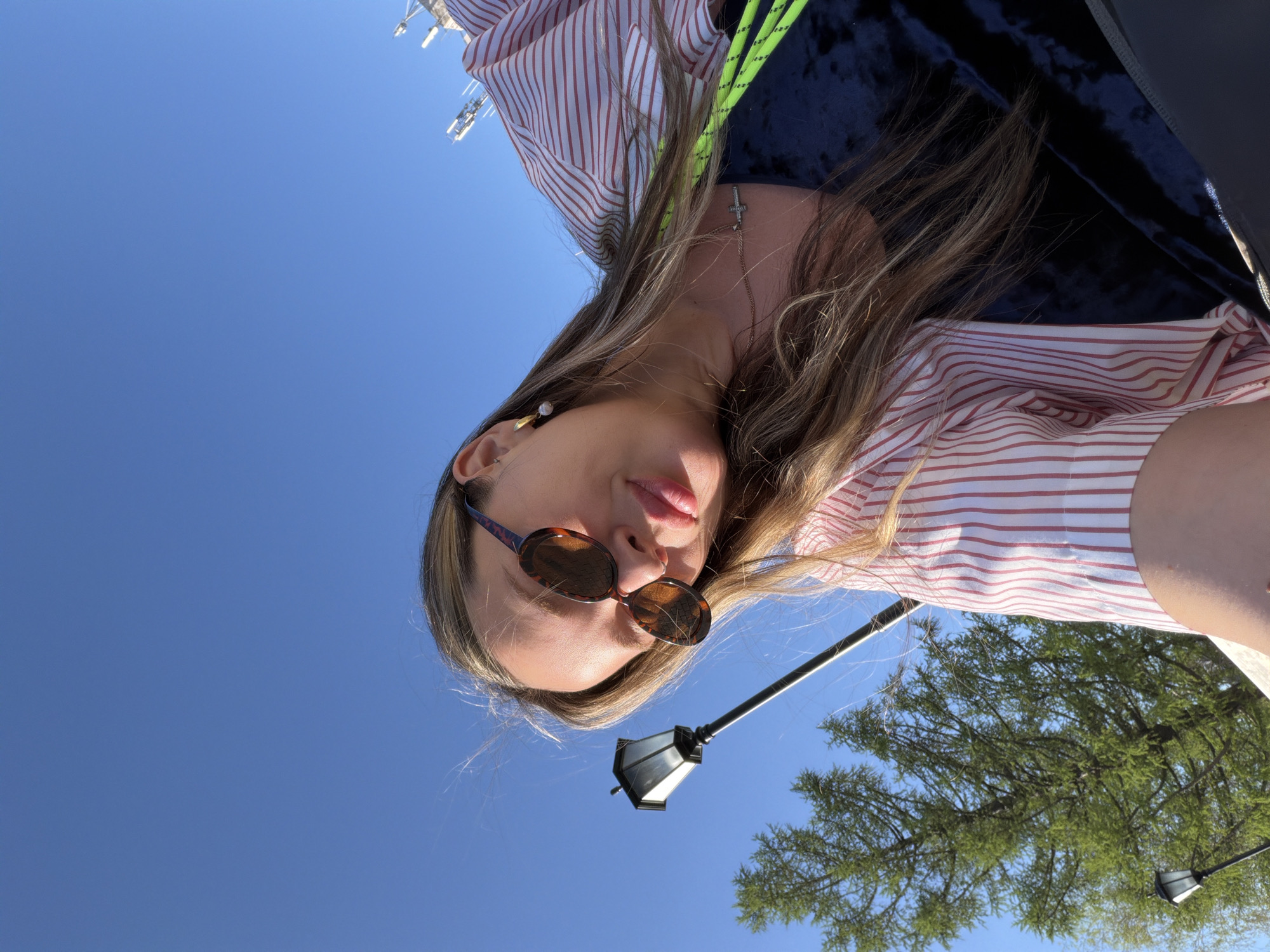 A woman wearing sunglasses and a red and white striped shirt is depicted in a low-angle selfie against a bright blue sky. Part of a tree and a street lamp are visible in the background. Due to the limited context and absence of clear landmarks, the specific location in Izhevsk, Russia cannot be ascertained.