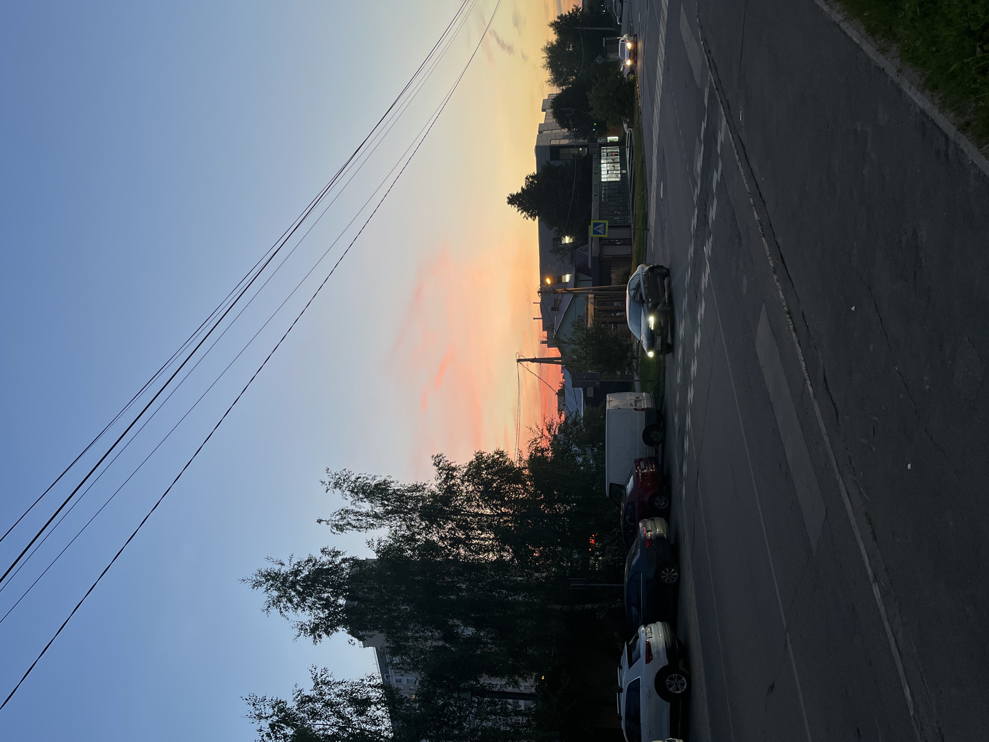 The image presents an urban street scene at dusk or early evening. The sky, transitioning from light blue overhead to vibrant orange and pink at the horizon, displays faint cloud formations. Several dark utility lines run diagonally across the upper portion of the frame.
On the left, an asphalt road features a series of parked vehicles: a white SUV, a dark sedan, a red sedan, and a white cargo van, positioned parallel to the curb. Behind these vehicles, a dense canopy of tall, dark-foliaged trees partially conceals a light-colored, multi-story building.
The central and right areas of the image are dominated by a two-lane asphalt road with a marked pedestrian crosswalk. A dark sedan with illuminated headlights is visible near the crosswalk, moving from left to right. Another vehicle with active headlights is discernible further down the road. Along the right side, commercial buildings are present, including a smaller, light-colored structure with a dark roof and an illuminated pedestrian crossing sign on a pole, and a larger building featuring a dark, reflective facade. Artificial lighting emanates from streetlights and building interiors. No individuals are visible. The setting is consistent with a city street environment during twilight hours.