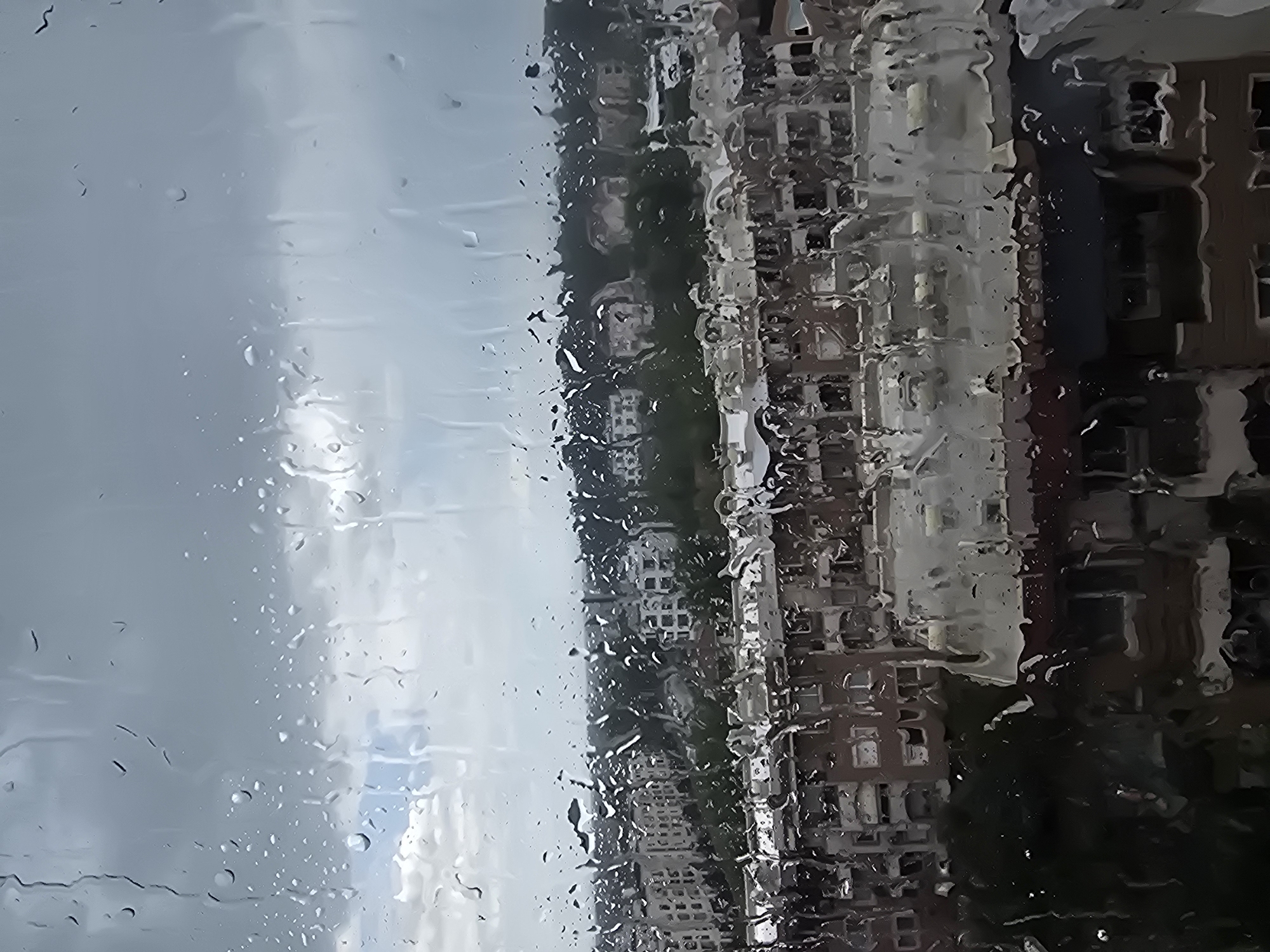 A photograph depicts an urban landscape viewed through a rain-streaked window. The sky is overcast, and the buildings across the street appear distorted due to the water droplets on the glass. The location is unspecified. The primary elements are the raindrops, the blurred cityscape, and the cloudy sky. There are no people visible in the frame. The image captures the atmosphere of a rainy day.