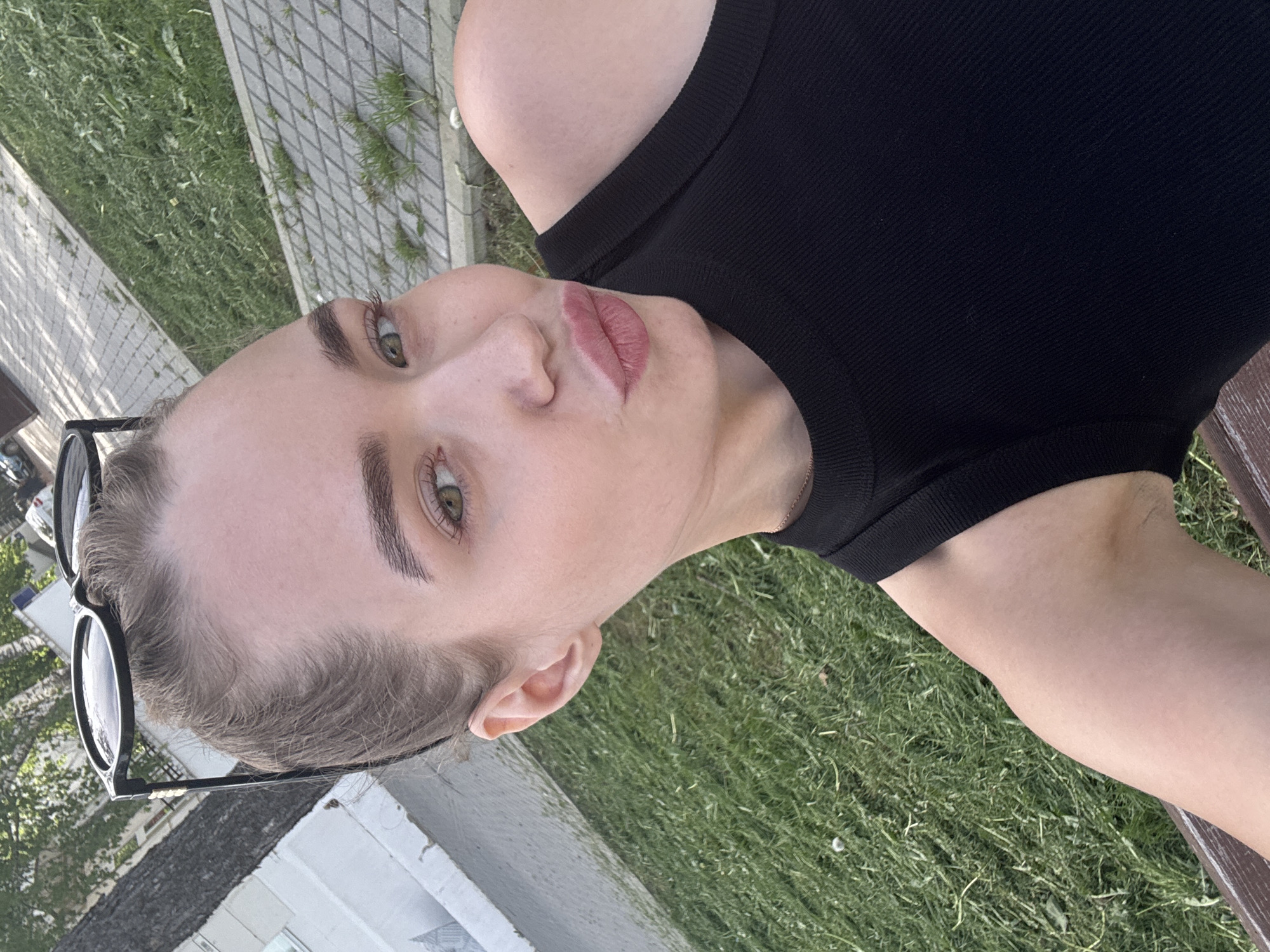 A woman is shown in an eye-level, close-up shot, likely taken on a bench outside, as suggested by the presence of grass. The setting might be Chaikovsky, Russia. She is wearing a black tank top, and sunglasses are perched on her head. She is facing the camera with her lips slightly puckered. There is a brick pavement visible in the background as well.