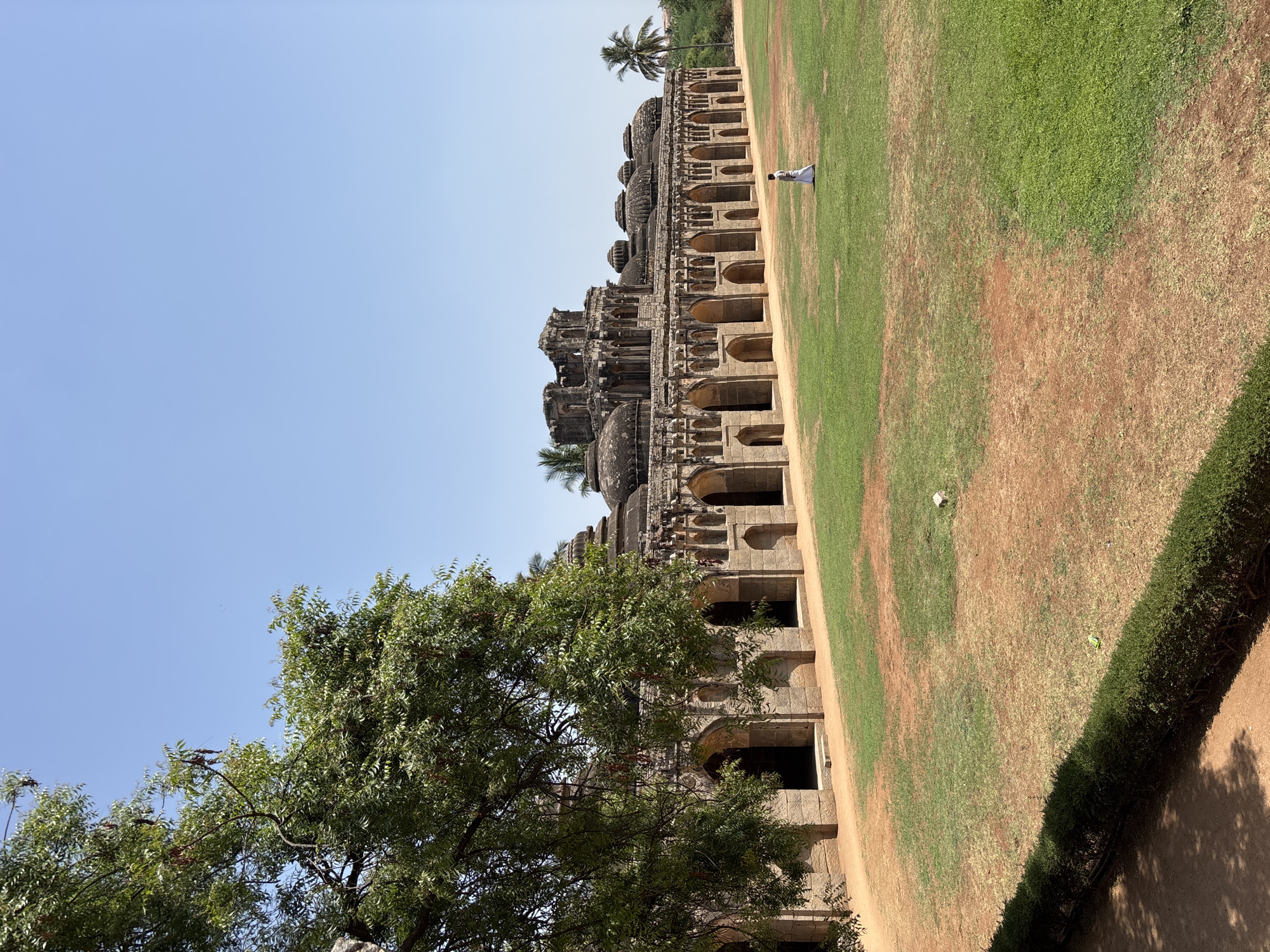 The image shows the Elephant Stables in the city of Kamalapura, India, a long, single-story building with multiple domed chambers. The structure is made of stone with arched openings. A person in white stands near the building. A large grassy area, transitioning from green to brown soil, stretches beside the stables. A tree is in the lower left corner. The sky is a clear blue.