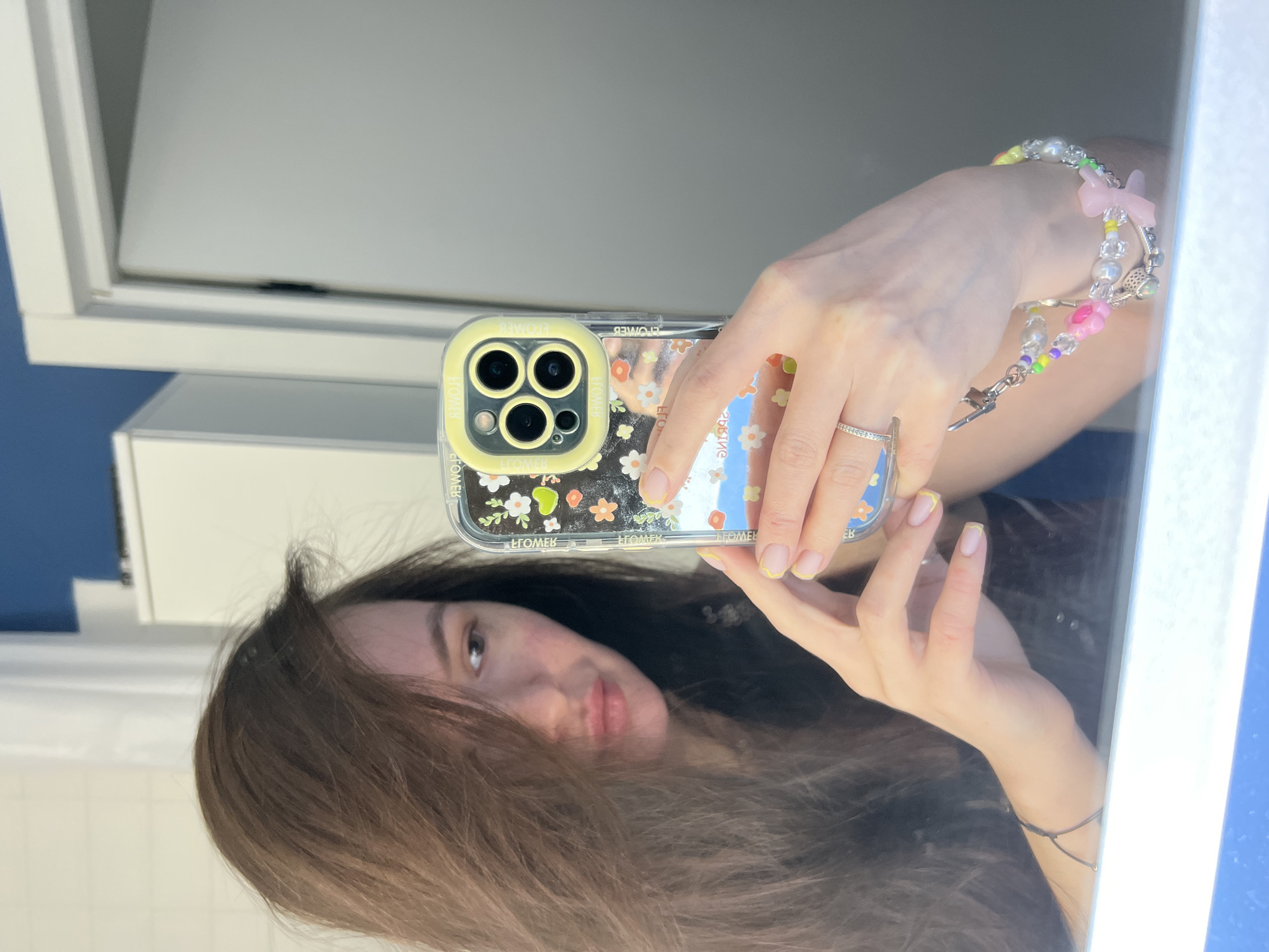 A person is taking a selfie in a bathroom with a phone. The phone is an iPhone with a clear case decorated with small flowers and a yellow border around the camera lenses. The person has long, brown hair. They are wearing a beaded bracelet with a charm on their wrist and a ring on their finger. The bathroom has white cabinets and a blue wall. The location is not identifiable as Kazan, Russia based on the provided information.