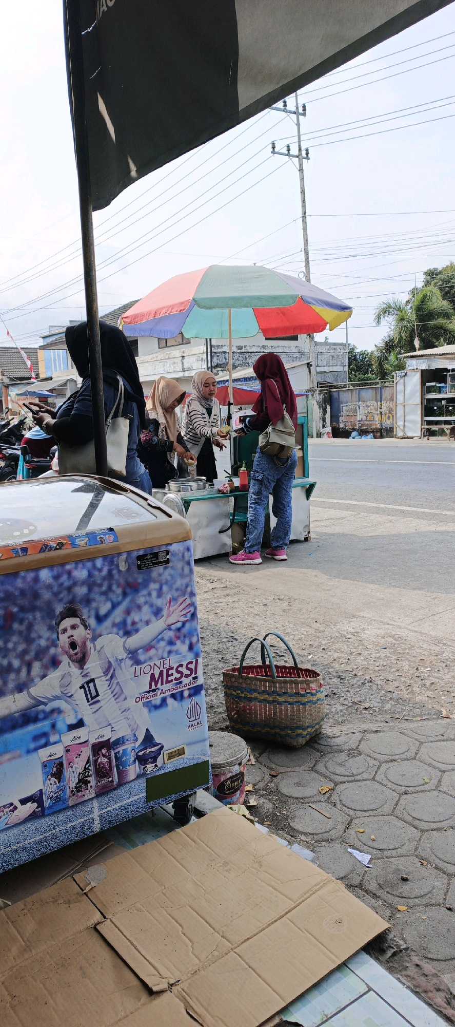 This is a vertical, eye-level shot of a street-side food stall in Kasiyan, Indonesia, featuring three women, a cooler, and a food cart under a colorful umbrella. In the foreground, a large cooler is adorned with a prominent image of Lionel Messi, a soccer player, and branding for "Alice" ice cream and "Halal Indonesia." To the right of the cooler is a woven basket with a red interior and blue and tan accents. Behind the cooler and basket, a food cart is set up under a multi-colored umbrella with red, yellow, green, and blue sections. Three women are engaged at the food cart. One woman on the left wears a dark hijab and a white top with dark stripes, holding a phone in her left hand. The woman next to her, in the center, wears a tan hijab and a black top. The third woman, on the right, wears a red hijab and a maroon jacket, holding a small cup and interacting with the woman in the tan hijab who appears to be serving something from the cart. The women are dressed in casual attire, with jeans being worn by two of them. The background shows a street with sparse traffic, buildings with tiled roofs, and a utility pole with multiple wires. The ground is paved with hexagonal concrete tiles and dirt. The overall scene depicts a typical street vendor operation in Indonesia.