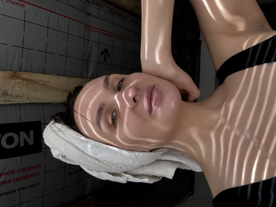 An image taken in the city of Chaikovsky, Russia, depicts a female individual in a close-up, head-and-shoulders portrait. Her head is wrapped in a white terry towel, with dark, wet hair visible along her forehead. Her left hand is positioned with fingers curled, resting against her jawline. She is wearing a dark, possibly black, garment, with a strap visible over her right shoulder. Distinct parallel strips of light and shadow are cast across her face, neck, and upper chest, originating from an unseen overhead source. The background reveals a dark grey, textured building material, consistent with a roofing underlayment or insulation membrane, featuring white grid lines, an arrow, and Cyrillic text. Visible Cyrillic phrases include "СТЫКОВ," "Лента для склеивания стыков" (Tape for gluing joints), and "ГОРЮЧИЙ МАТЕРИАЛ" (Flammable Material). A roughly cut wooden beam is present in the upper right portion of the background. The scene suggests an interior or semi-enclosed space, potentially under construction or within an attic.