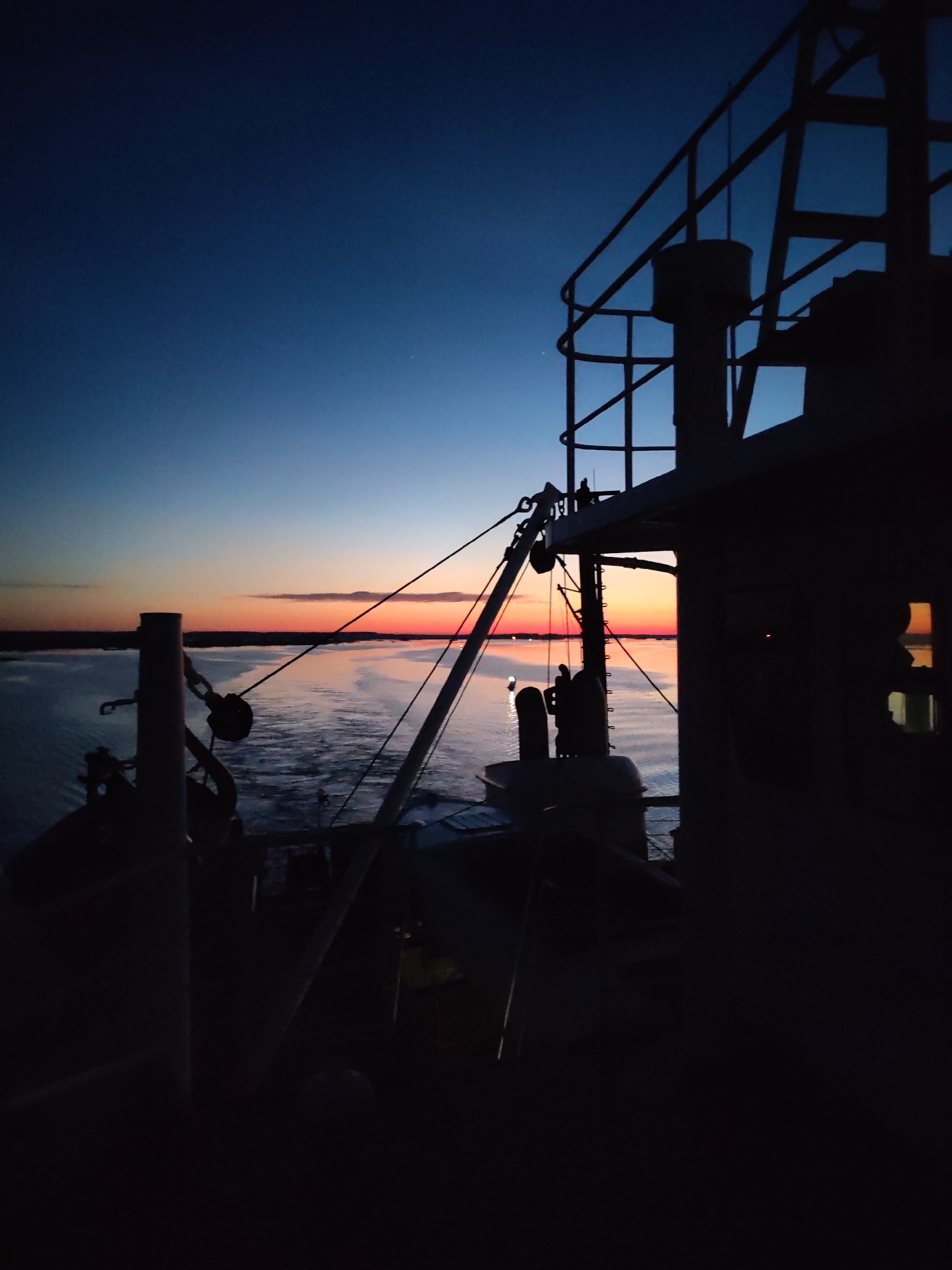 The image depicts a sunset viewed from the deck of a ship at sea. The sky transitions from dark blue at the top to orange and red near the horizon. The water reflects the colors of the sunset. The silhouette of ship's equipment, including railings, cables, and machinery, is prominent in the foreground and right side of the image. A light or buoy is visible on the water in the distance. The location cannot be determined as Bereg, Russia based on visual information in the image.