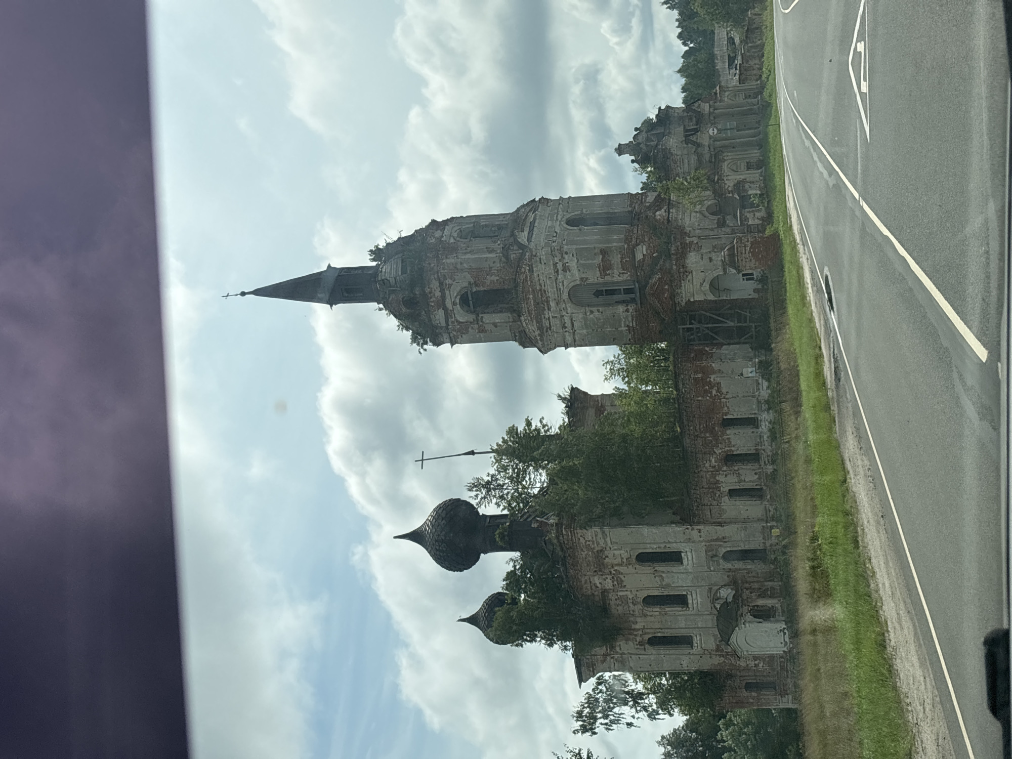 The image shows an abandoned, dilapidated church complex situated in Pryamik, Russia. The architecture features multiple domes and a tall bell tower, all in a state of severe disrepair with crumbling brickwork and vegetation growing over the structures. The church is located adjacent to a paved road, with a grassy verge between the road and the building. The sky is partly cloudy, with cumulus clouds against a light blue background. The scene appears to be captured from a vehicle, as indicated by the darkened upper portion of the image, possibly a glare or tint on a car window. There are no people visible in the image.